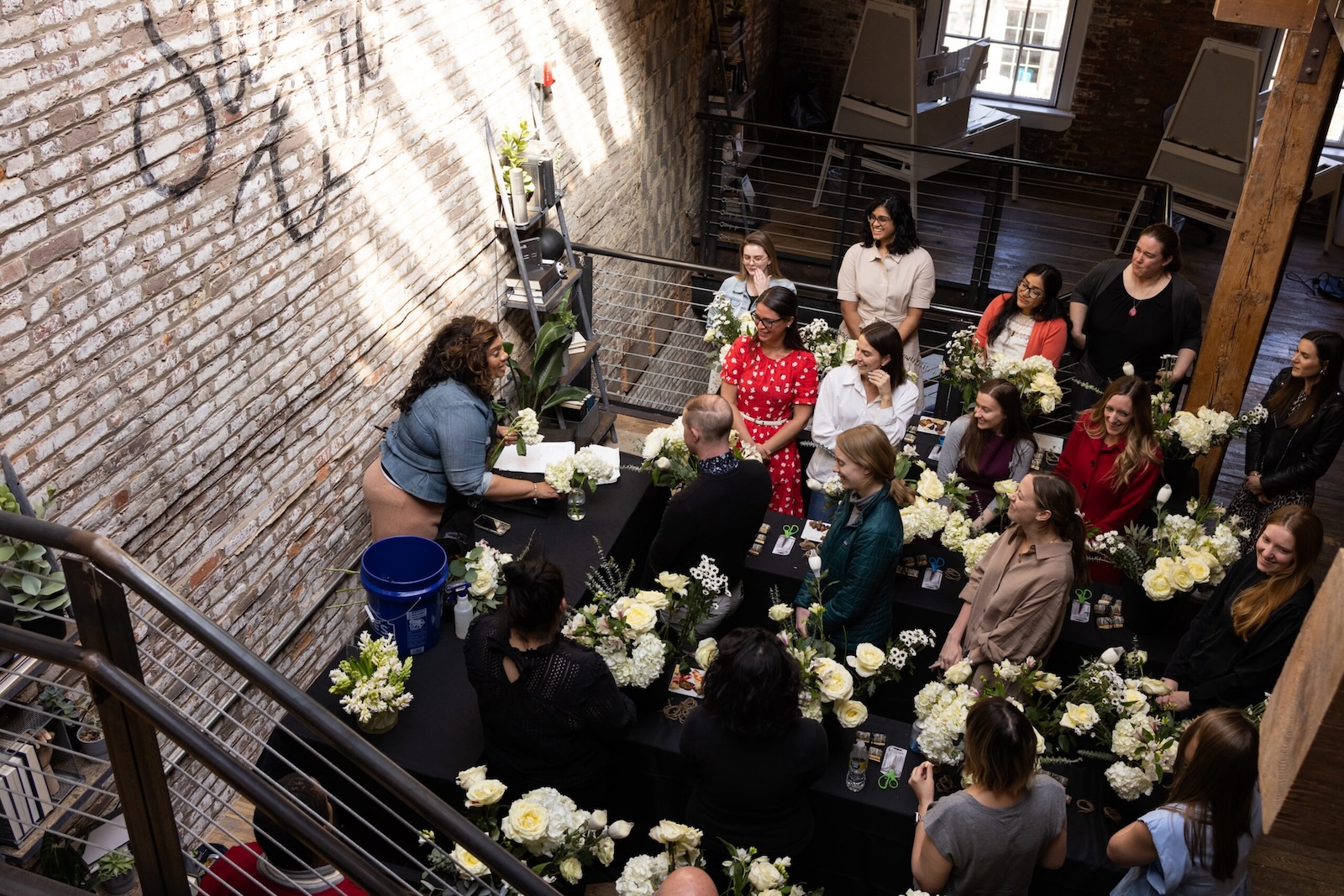 A group of people are gathered in a brick-walled room with large windows. They are attending a flower arranging workshop led by a person at the front, surrounded by white and yellow flowers. They are all focused on the demonstration, with flowers spread out on the tables.