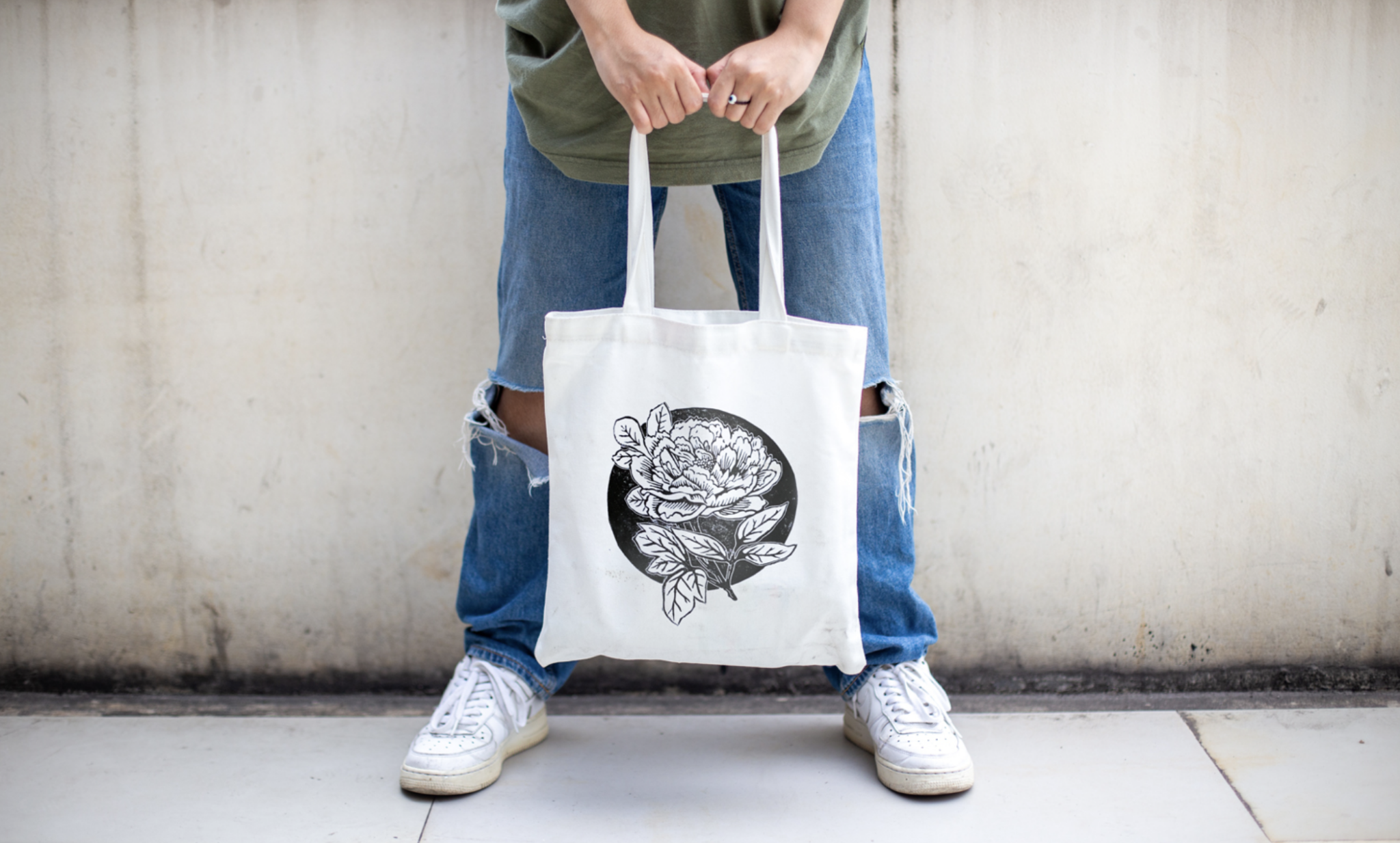 Person wearing ripped blue jeans and white sneakers holding a white canvas tote bag featuring a black floral design. The background is a plain, textured off-white wall, and the focus is on the tote bag and the lower half of the person’s body.