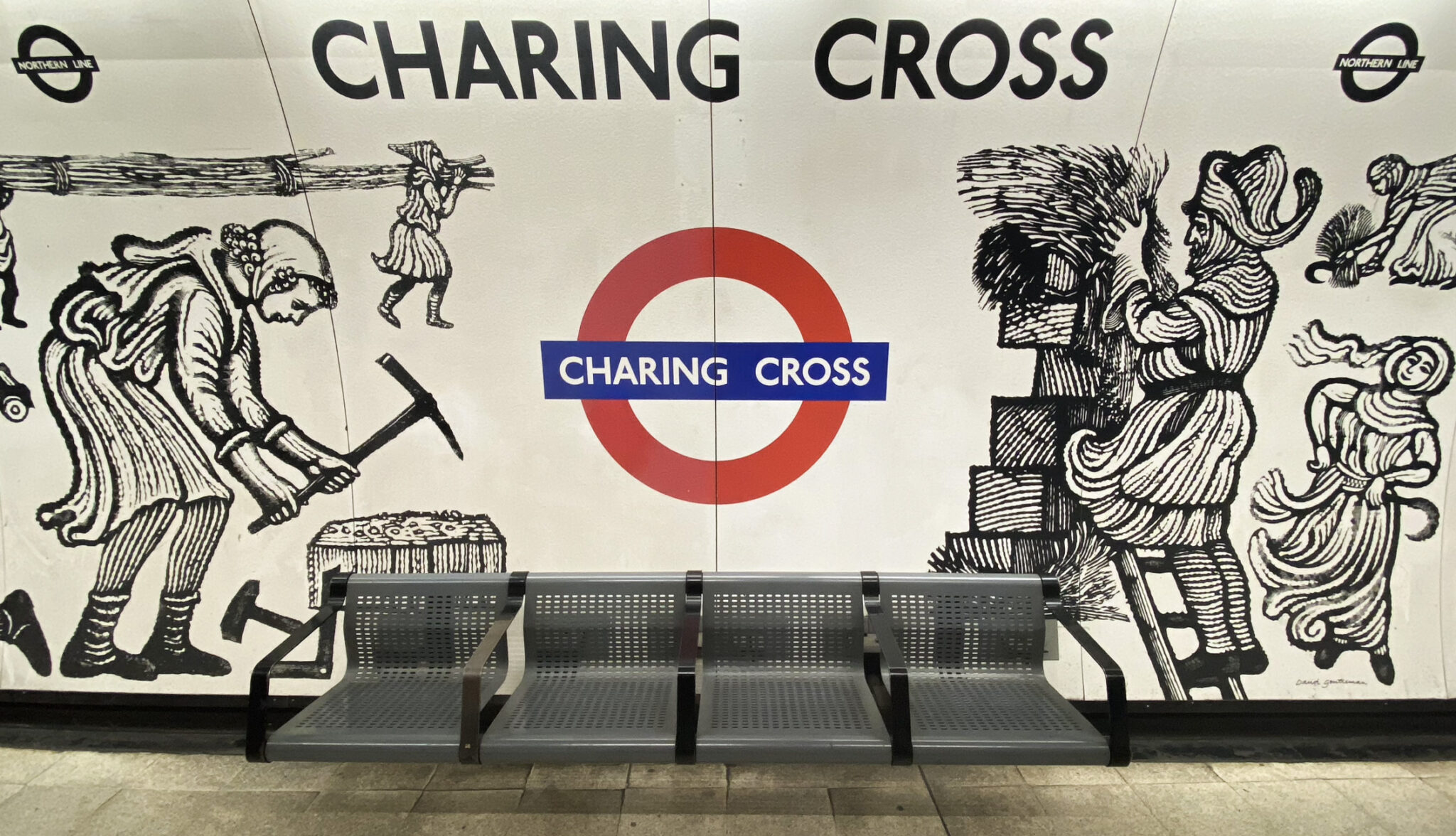 A section of a subway station wall at Charing Cross features black and white historical illustrations, a red and blue roundel with "Charing Cross" in the center, and three perforated metal benches in the foreground.