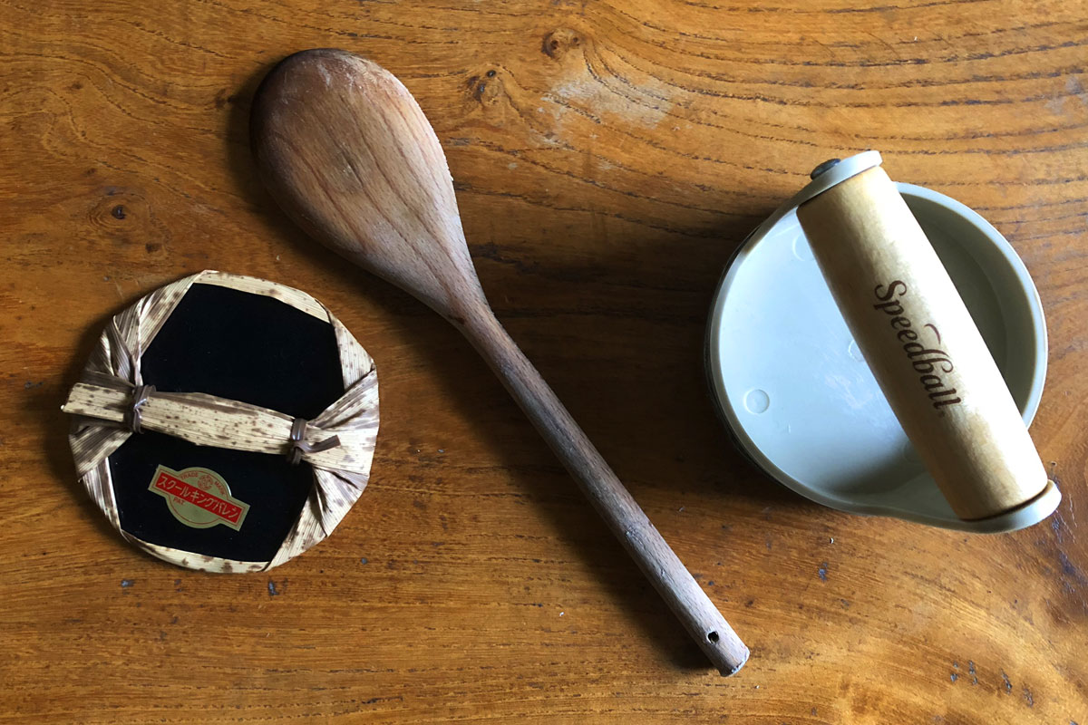 A wooden spoon, a ceramic pot with a lid marked "Speedball," and a wrapped item with a red label sit on a wooden surface. The spoon has a light brown hue, while the pot and item create a rustic kitchen atmosphere.