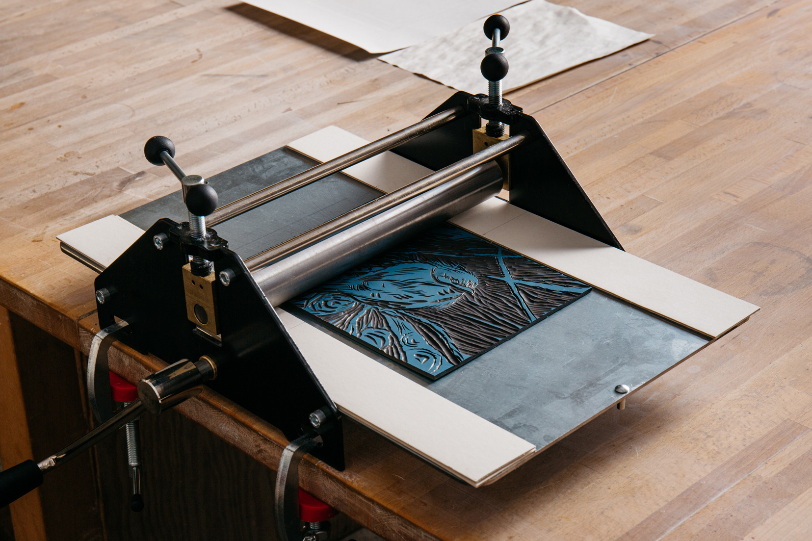 A black etching press on a wooden table, holding a blue inked linocut print. A sheet of paper partially covers the print, ready to be rolled through the press. The surrounding area is neatly organized, with additional paper in the background.
