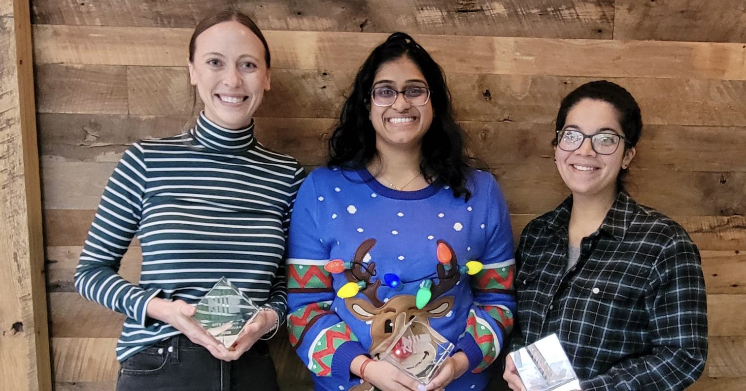Three people are standing side by side, smiling and holding awards. The person on the left wears a striped turtleneck, the person in the center is in a festive reindeer sweater, and the person on the right wears glasses and a plaid shirt. The background features a wooden wall.