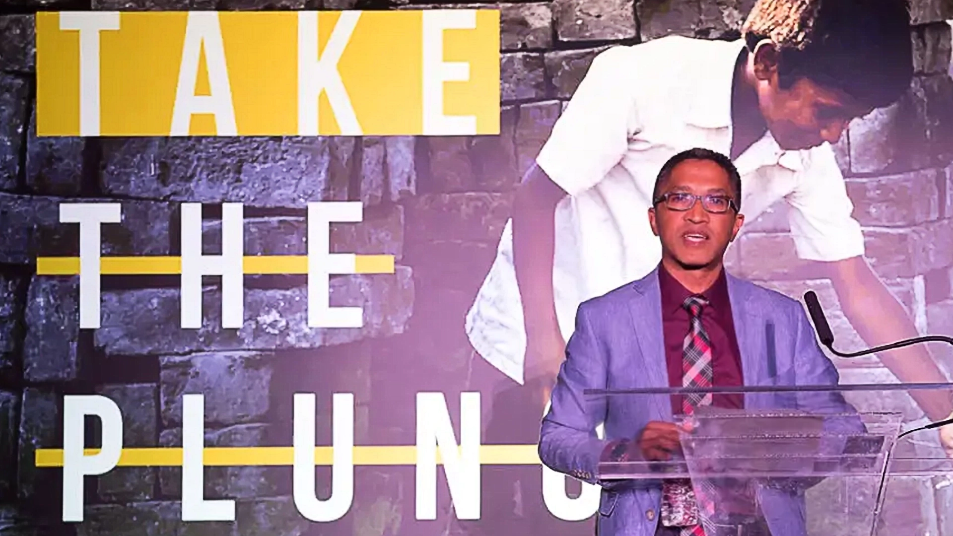 A man in a suit and tie stands at a podium speaking at the DefeatDD Event, with a backdrop displaying "TAKE THE PLUNGE" beside an image of a child leaning forward against a stone wall. This interactive event promises an engaging blend of dialogue and experiential marketing.