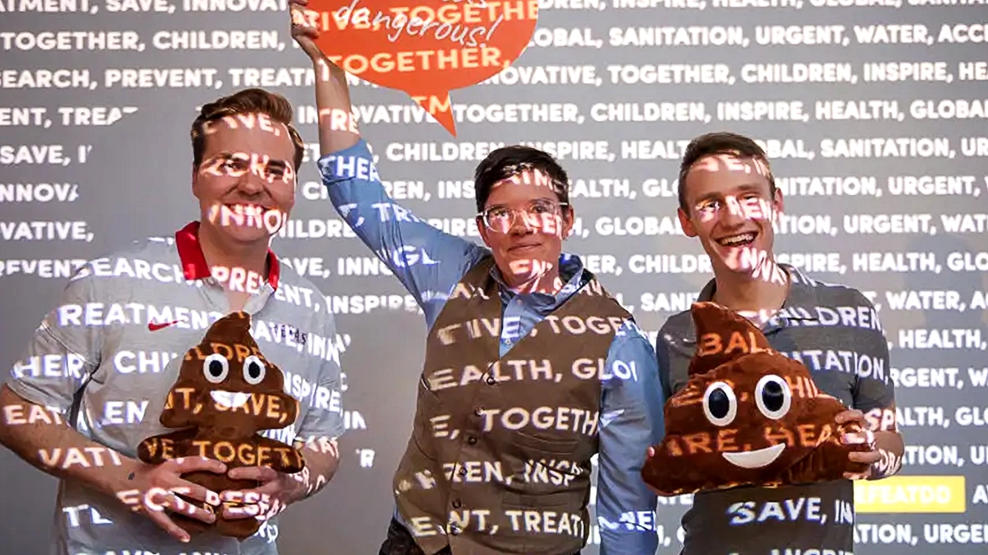 Three people smiling, two holding emoji poop pillows. Background projection includes words like "innovate," "health," and "together." A speech bubble above reads, "Together." The scene conveys a fun, collaborative atmosphere of health advocacy through experiential marketing.