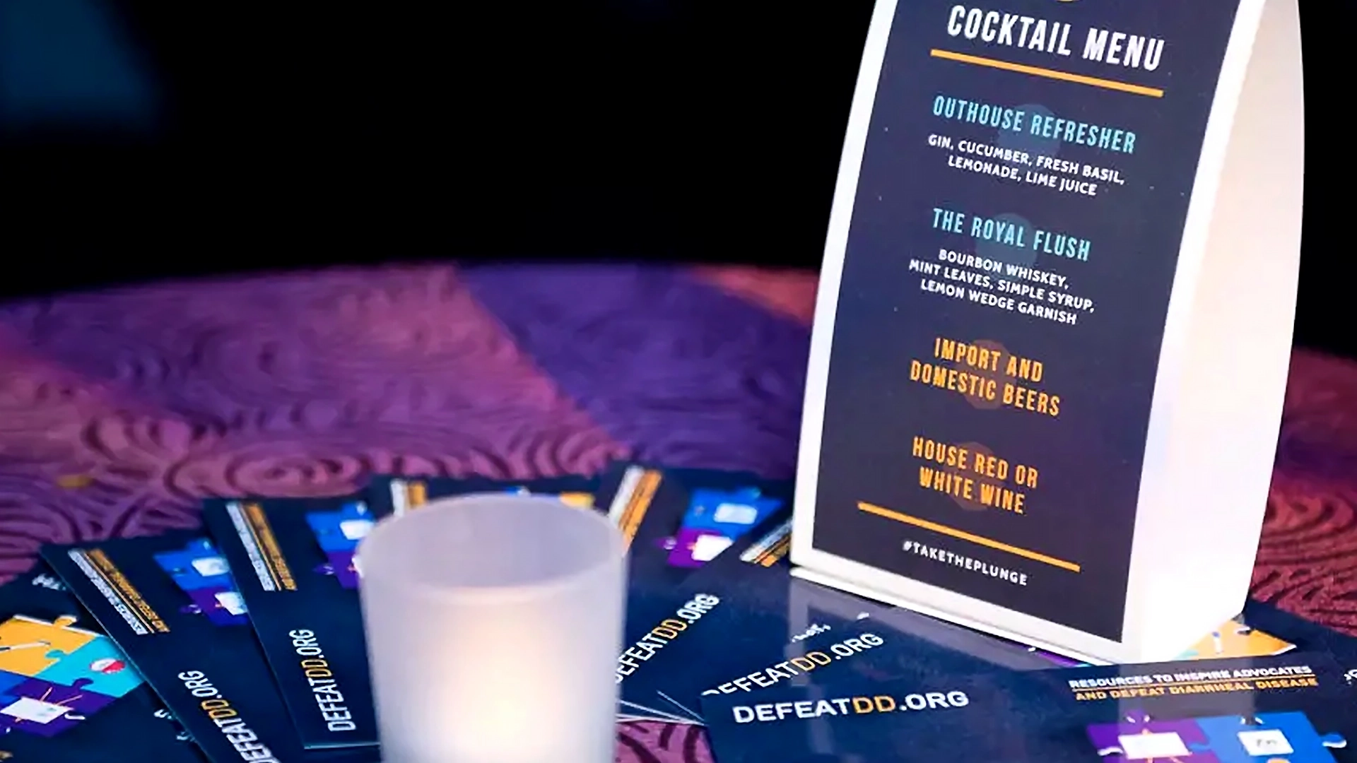 A cocktail menu on the table is surrounded by brochures promoting DefeatDD.org at an experiential event. A small, unlit candle sits in the foreground. The menu lists drinks like "Outhouse Refresher" and "The Royal Flush." The tablecloth features a dark, textured pattern.