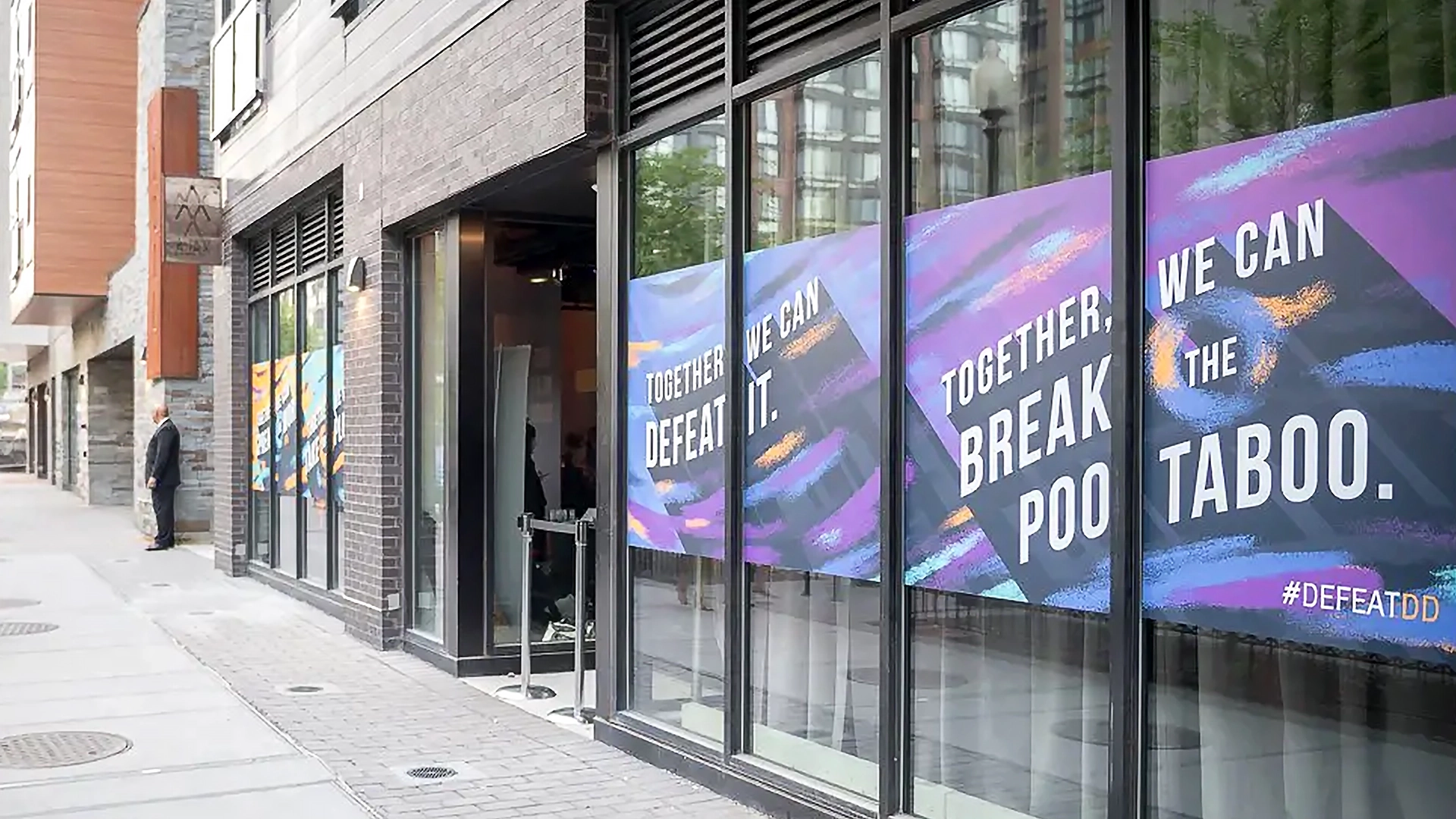 Street view of a building with large glass windows showcasing colorful signs that proclaim, "Together, we can defeat it. Together, we can break the taboo." A person is visible in the distance, hinting at an upcoming DefeatDD Event promoting health advocacy.