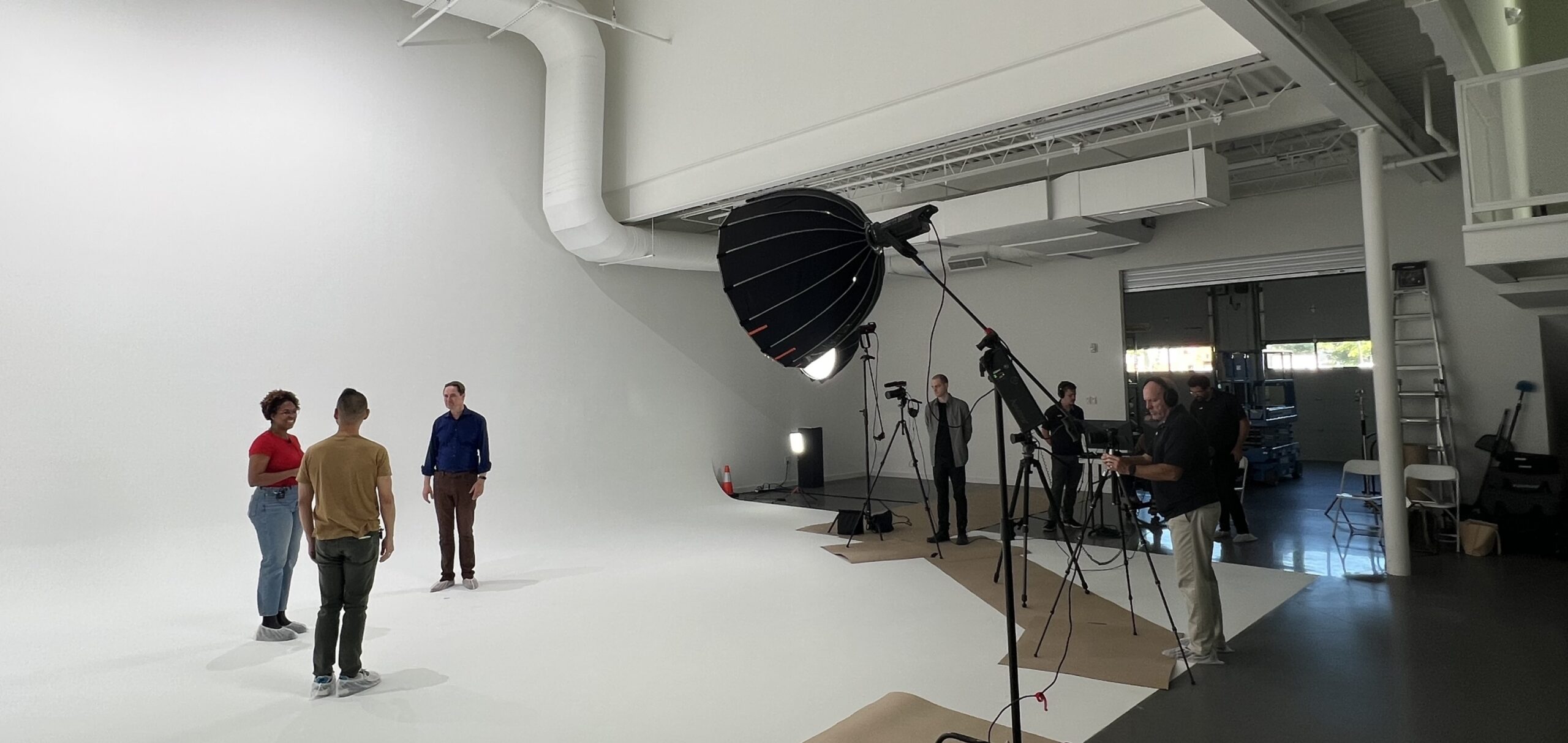 People are in a spacious studio with white walls and floor, involved in a photoshoot or video production. Some individuals are operating cameras and equipment, while two people stand under a large lighting umbrella. Various gear and backdrops are visible around.