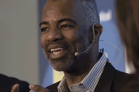 A man with short, graying hair, a beard, and a mustache is speaking about scientific research, wearing a checkered shirt and a blazer. He has a headset microphone on his cheek. The background is out of focus and features blue and yellow hues.