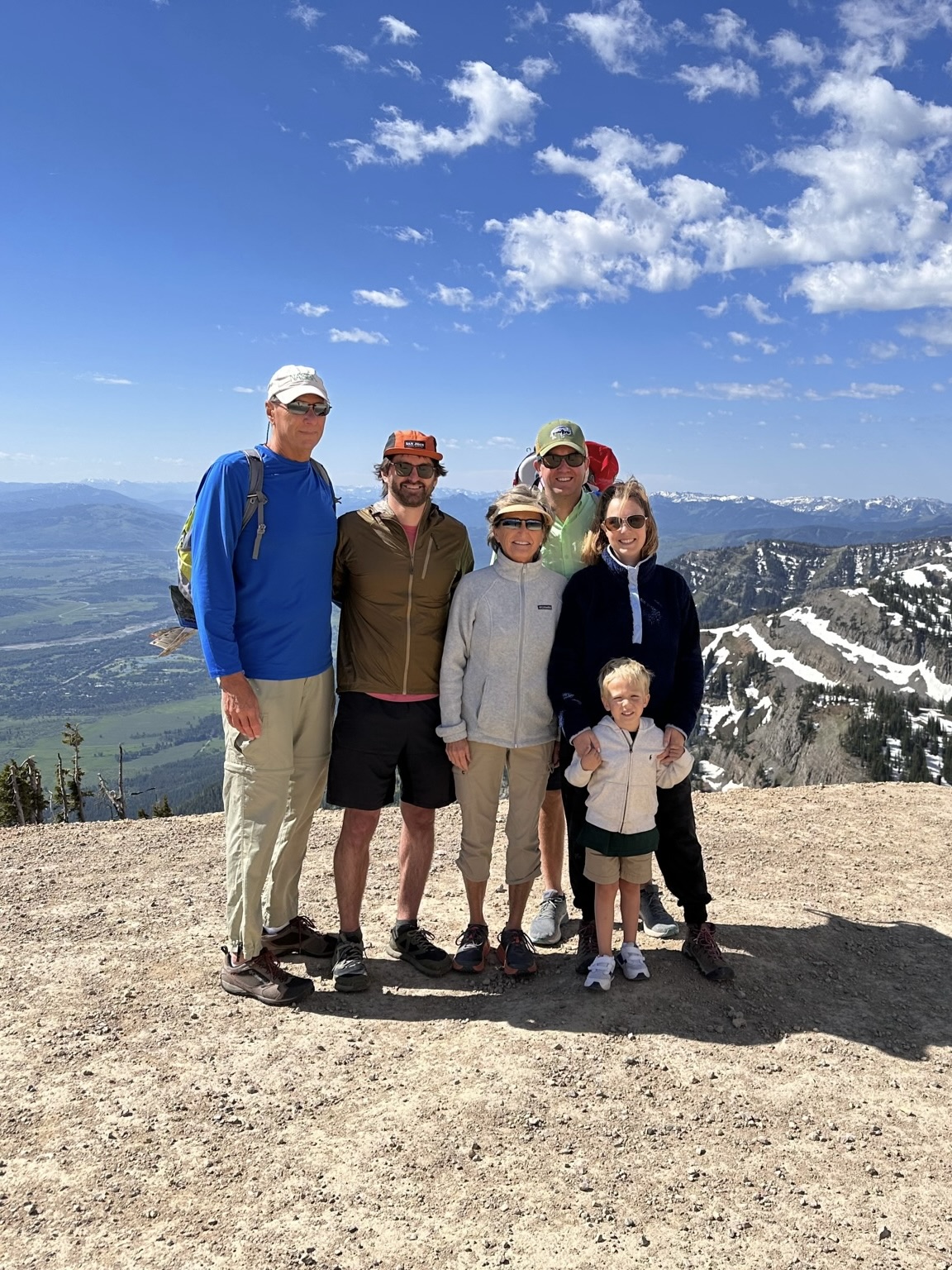 Six people, including a child, stand on a scenic mountain overlook with a vast landscape of valleys and snow-capped peaks in the background. They are dressed in outdoor clothing, smiling, and standing close together under a clear blue sky.