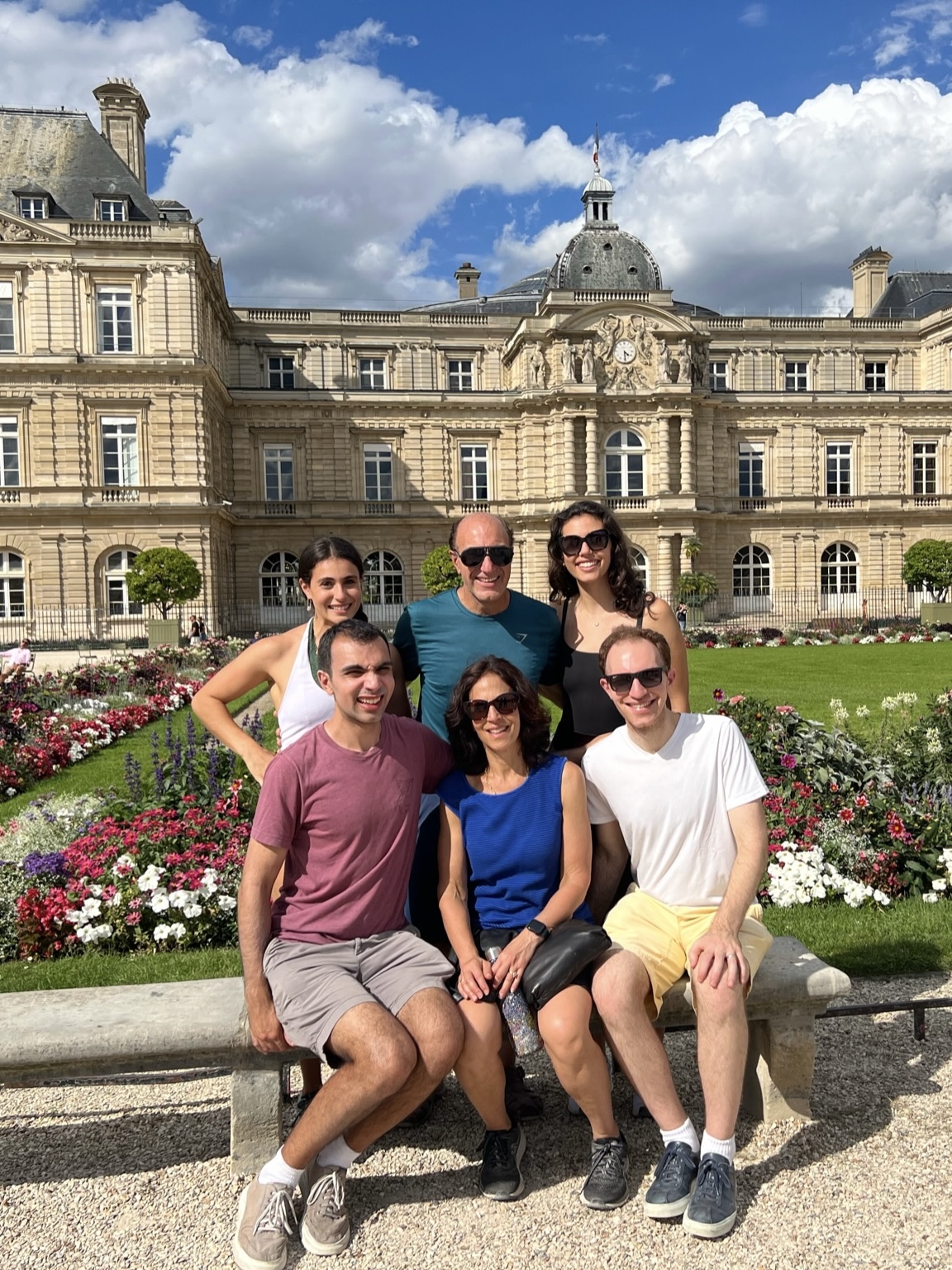 A group of six people posing and smiling in front of the Luxembourg Palace in Paris on a sunny day, clearly enjoying their employee vacation benefits. Three people stand in the back while the other three sit on a bench in front. Flower gardens and the historic building are visible in the background.