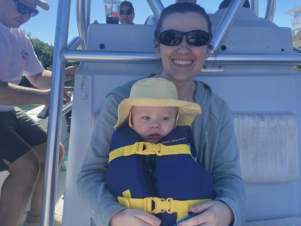 A woman wearing sunglasses and smiling holds a baby in a blue and yellow lifejacket and yellow hat on a boat, clearly taking advantage of her employee travel benefits. Two other people are visible behind them, enjoying the sunny day.