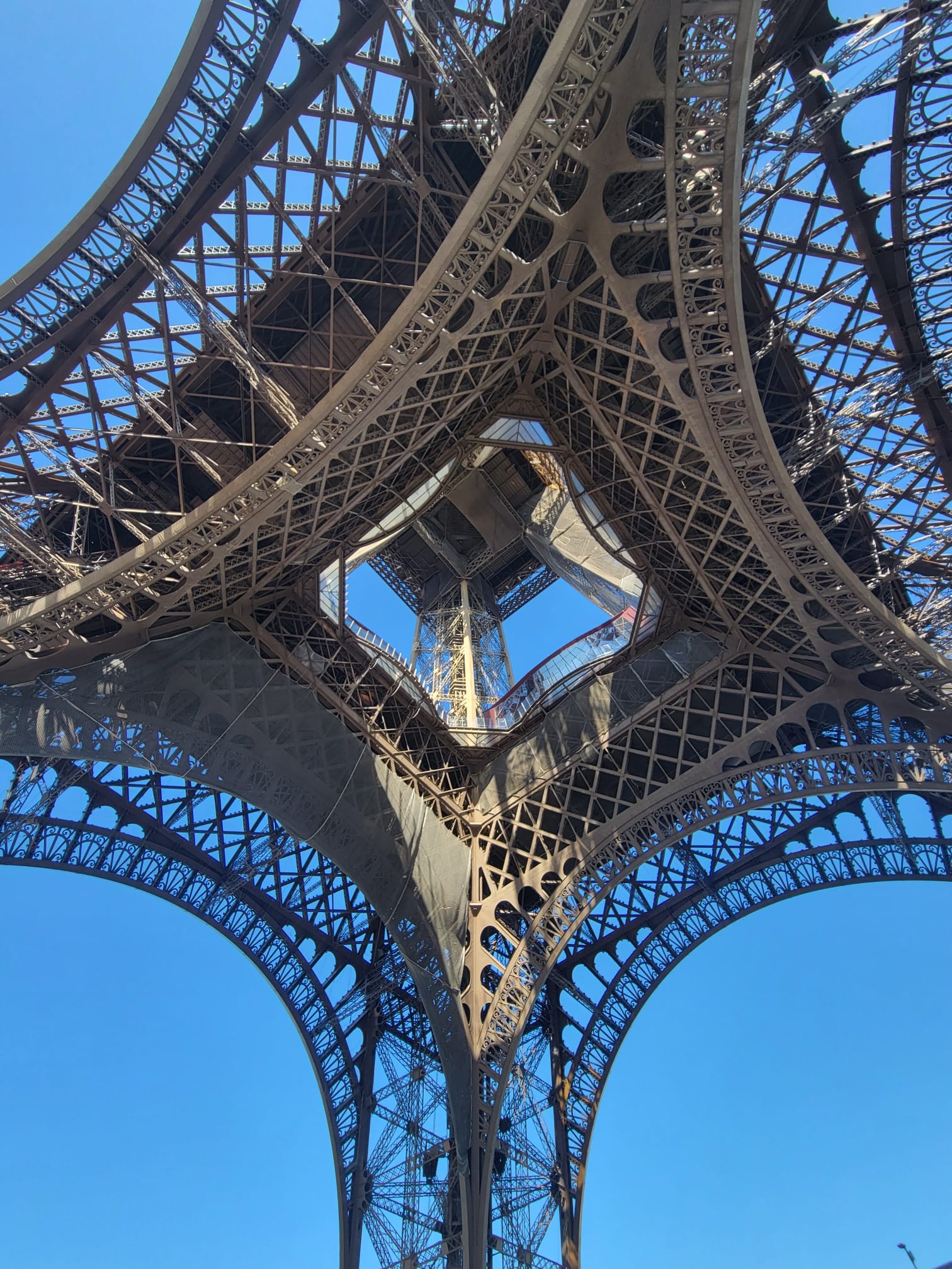 A view looking up through the intricate iron latticework of the Eiffel Tower, showcasing its graceful arches and geometric patterns set against a clear blue sky.
