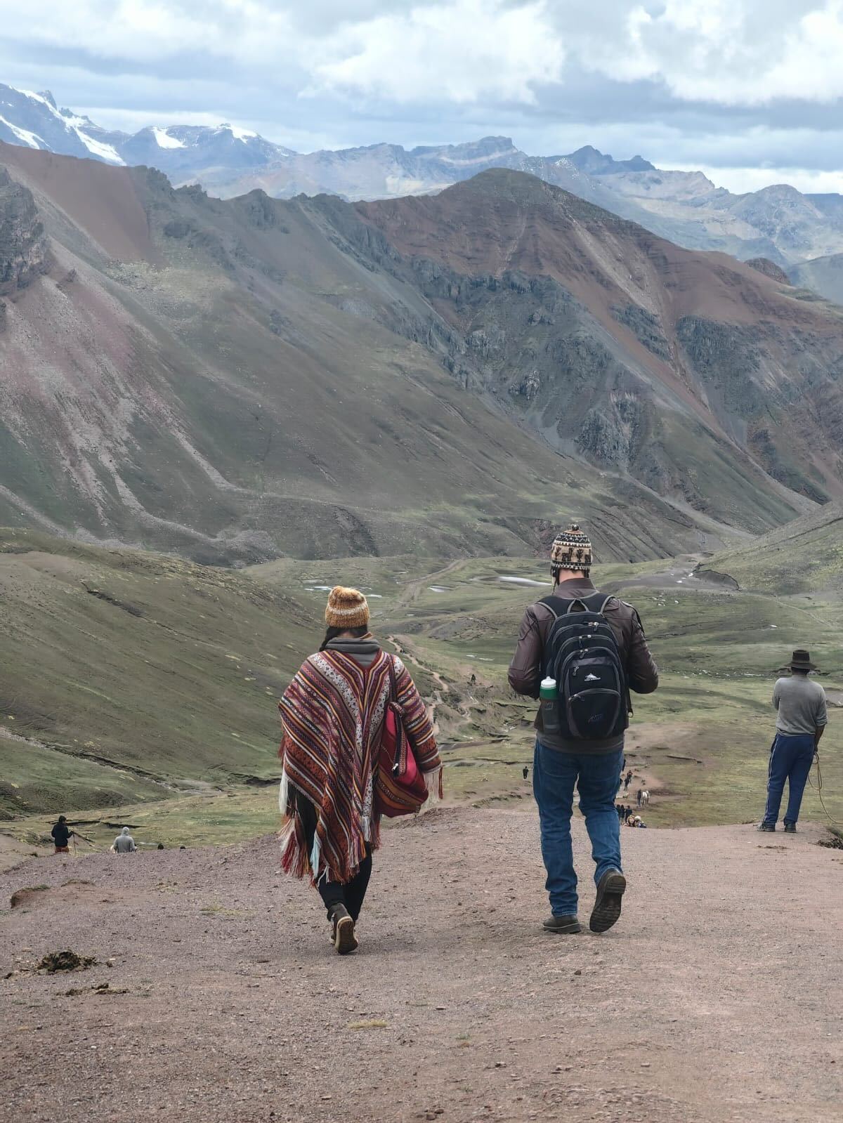 Two people dressed in warm clothing and hats walk down a mountain trail with rocky, green terrain and high peaks in the background. The person on the left wears a colorful shawl, while the person on the right carries a blue backpack. Other hikers are seen in the distance.