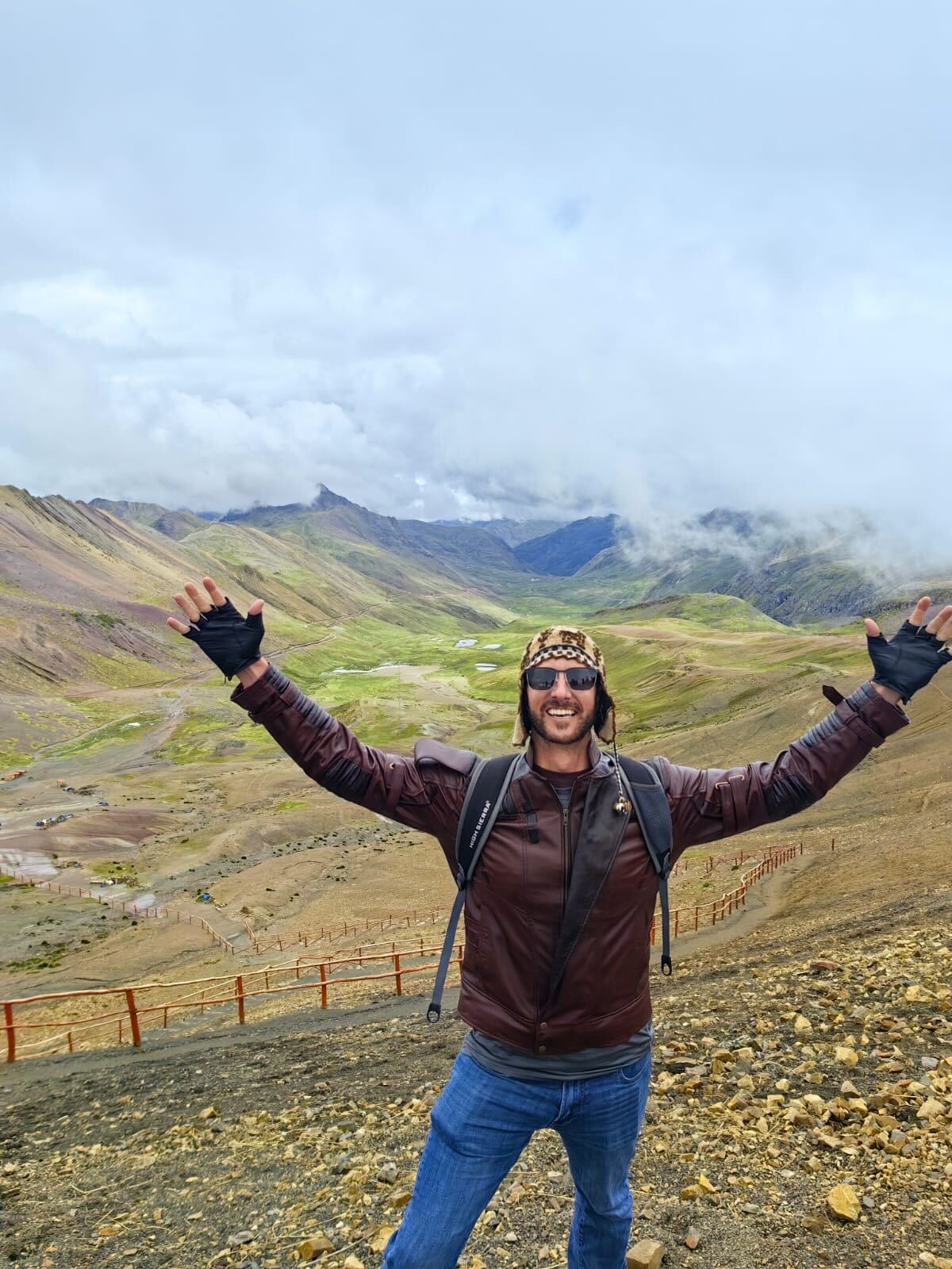 A person in a brown jacket and blue jeans stands on a mountainous trail with arms raised in joy, showcasing the excitement of employee travel. They wear gloves, a headband, sunglasses, and a backpack. The backdrop features rolling hills, scattered rocks, a lush green valley, and a cloud-filled sky.