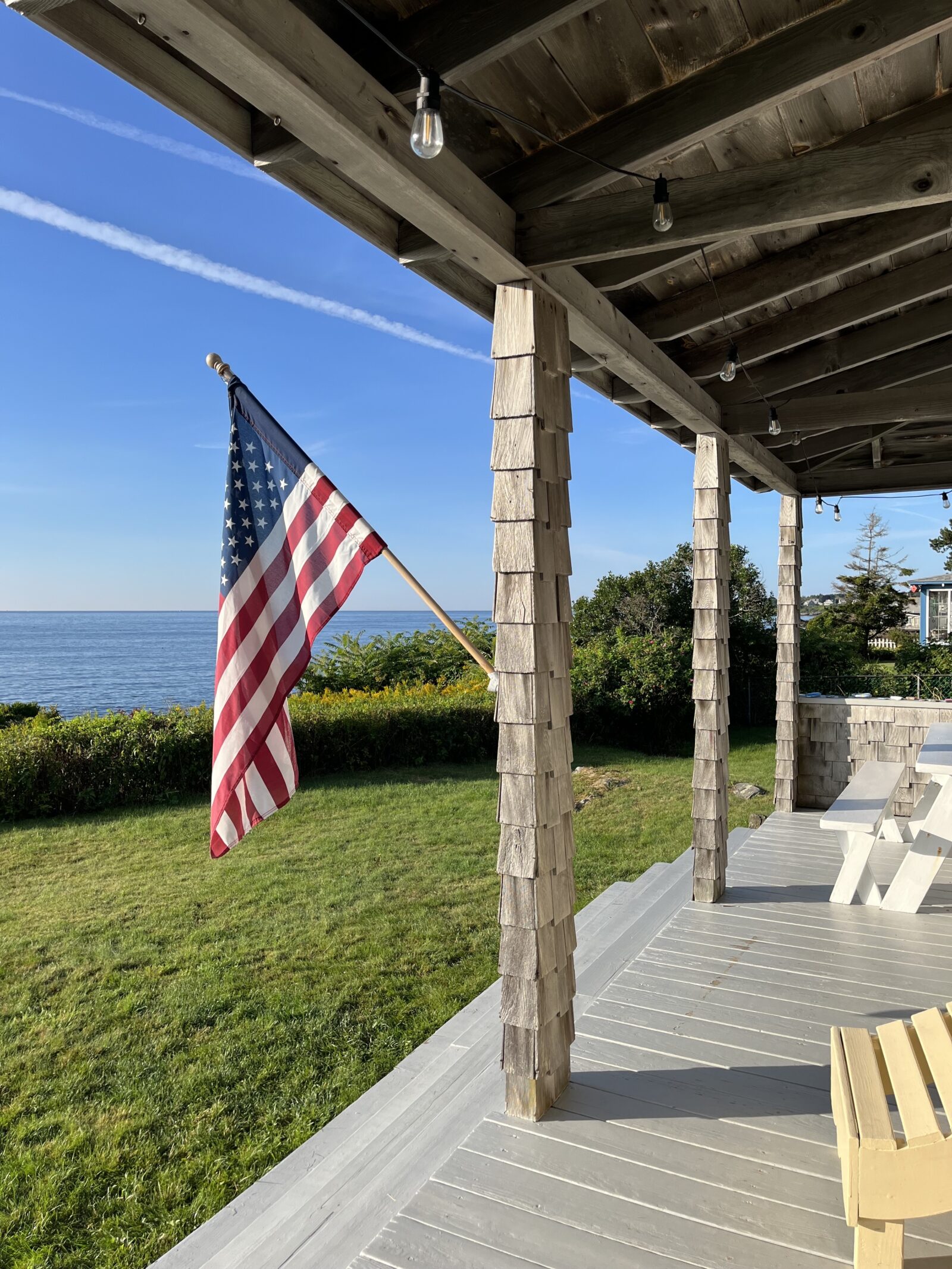 A wooden porch with shingled pillars overlooks a grassy yard and a calm sea under a clear blue sky. An American flag hangs from one of the pillars. The porch is partially shaded under the roof, with string lights hanging above.