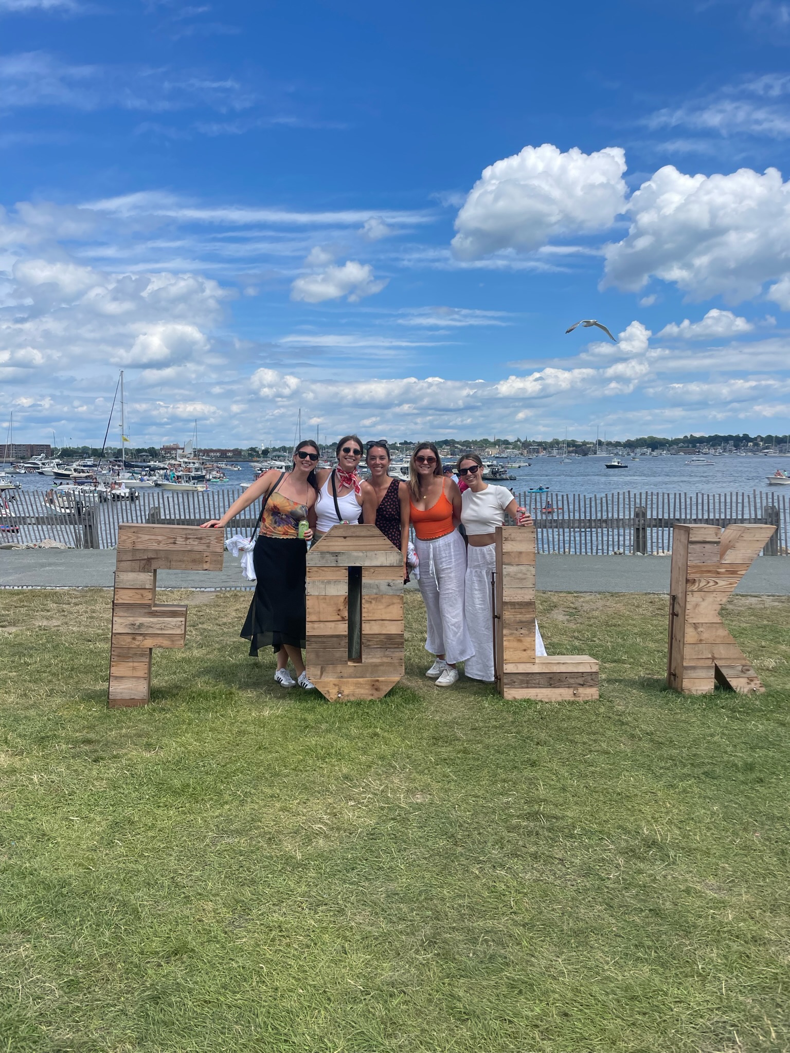A group of five people stands together outdoors, smiling at the camera, with their arms around one another. They are posing next to large wooden letters spelling "FOLK." The background features a scenic view of water, boats, and a partly cloudy sky.