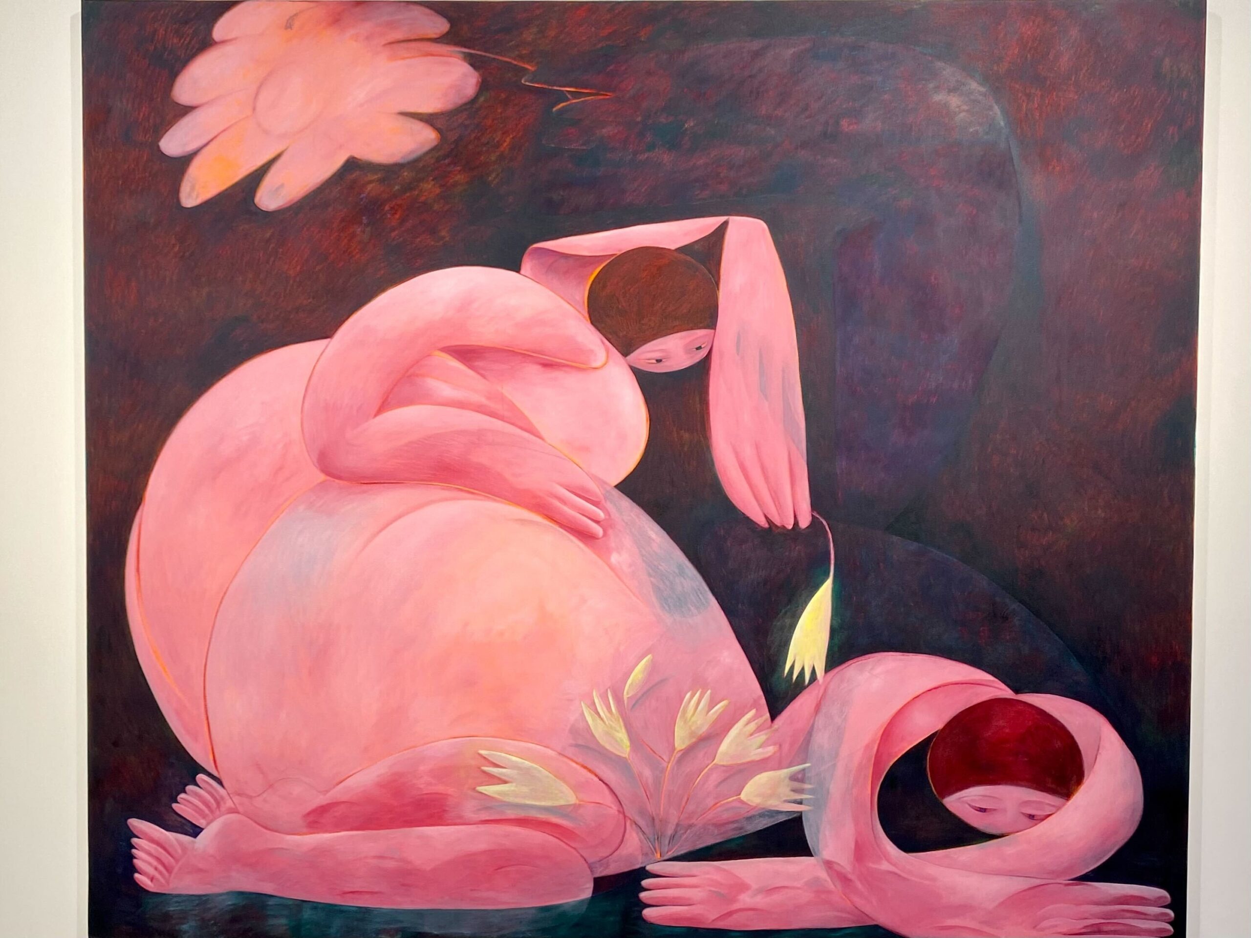 Abstract painting featuring two pink human-like figures against a dark background. One figure is seated, cradling a flower, while the other figure is bent over with a contemplative posture. Both have rounded, flowing forms, and the scene evokes a surreal, dream-like atmosphere.