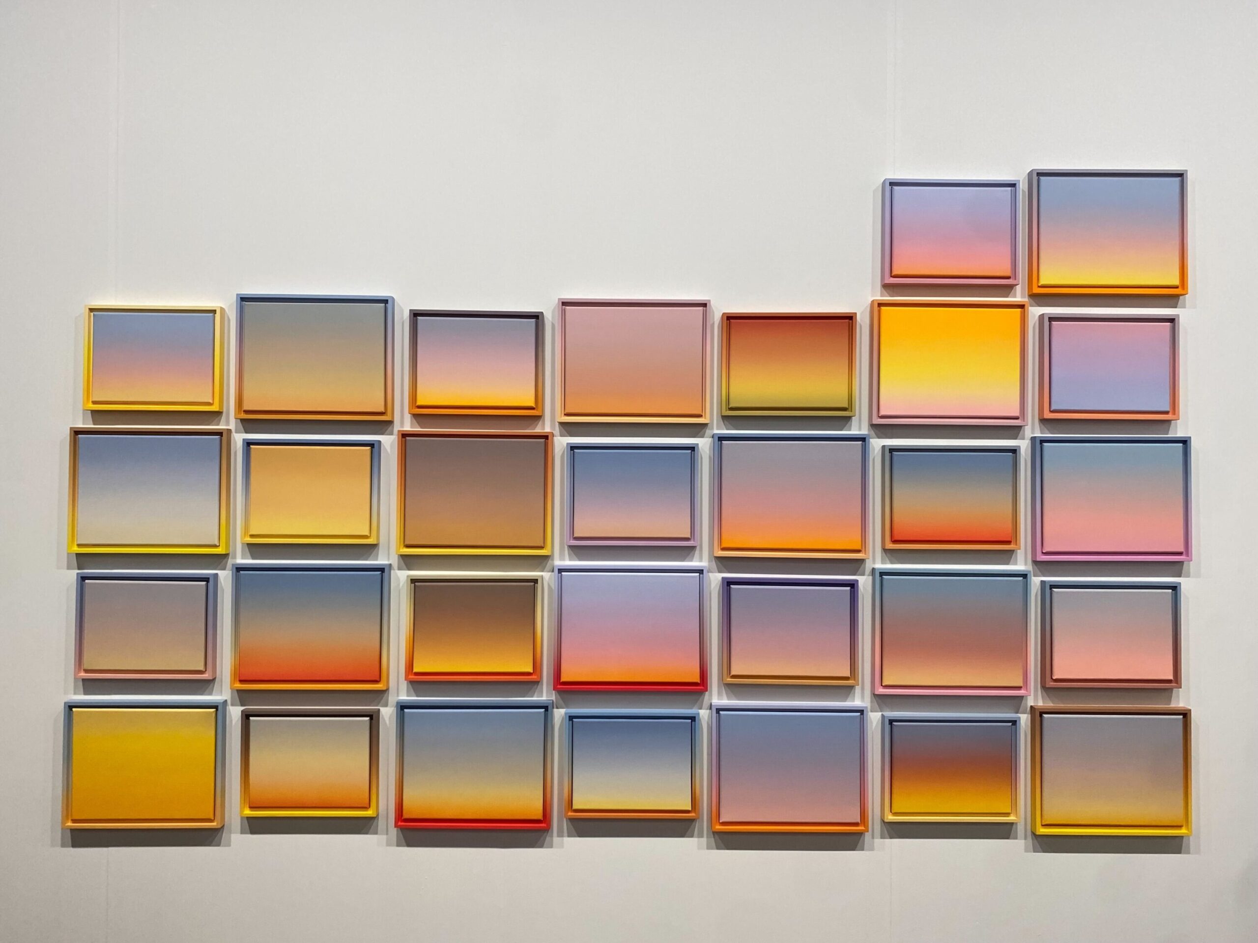 A wall display of 25 framed square pieces, each featuring a gradient of warm colors like yellow, orange, pink, and purple. The arrangement forms an irregular grid pattern, creating a visually striking mosaic effect.