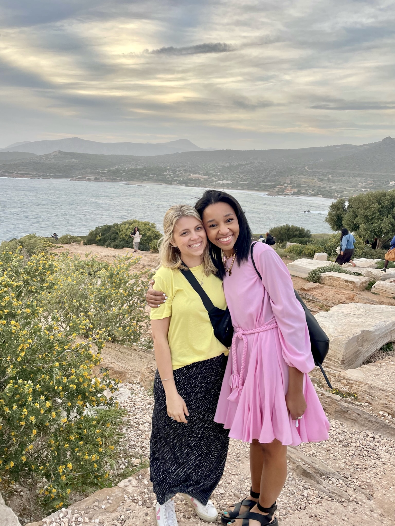 Two women stand closely, smiling at the camera against a scenic backdrop of a coastal landscape with mountains, water, and cloudy skies. One wears a yellow top and black skirt, and the other wears a light pink dress. Wildflowers and greenery are visible in the foreground.