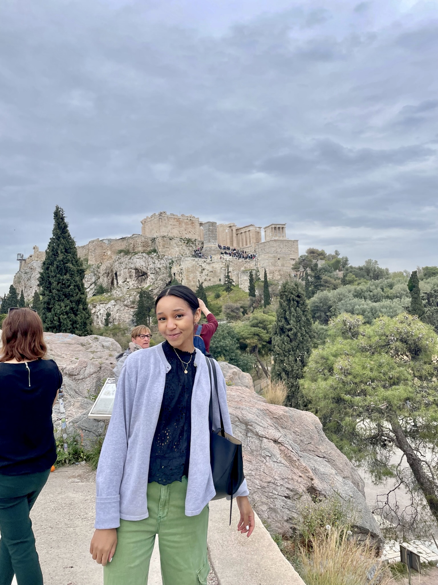 A person in a blue jacket, black shirt, and green pants stands smiling in front of a viewpoint with the Acropolis of Athens in the background. Other visitors and lush greenery are visible under a cloudy sky.