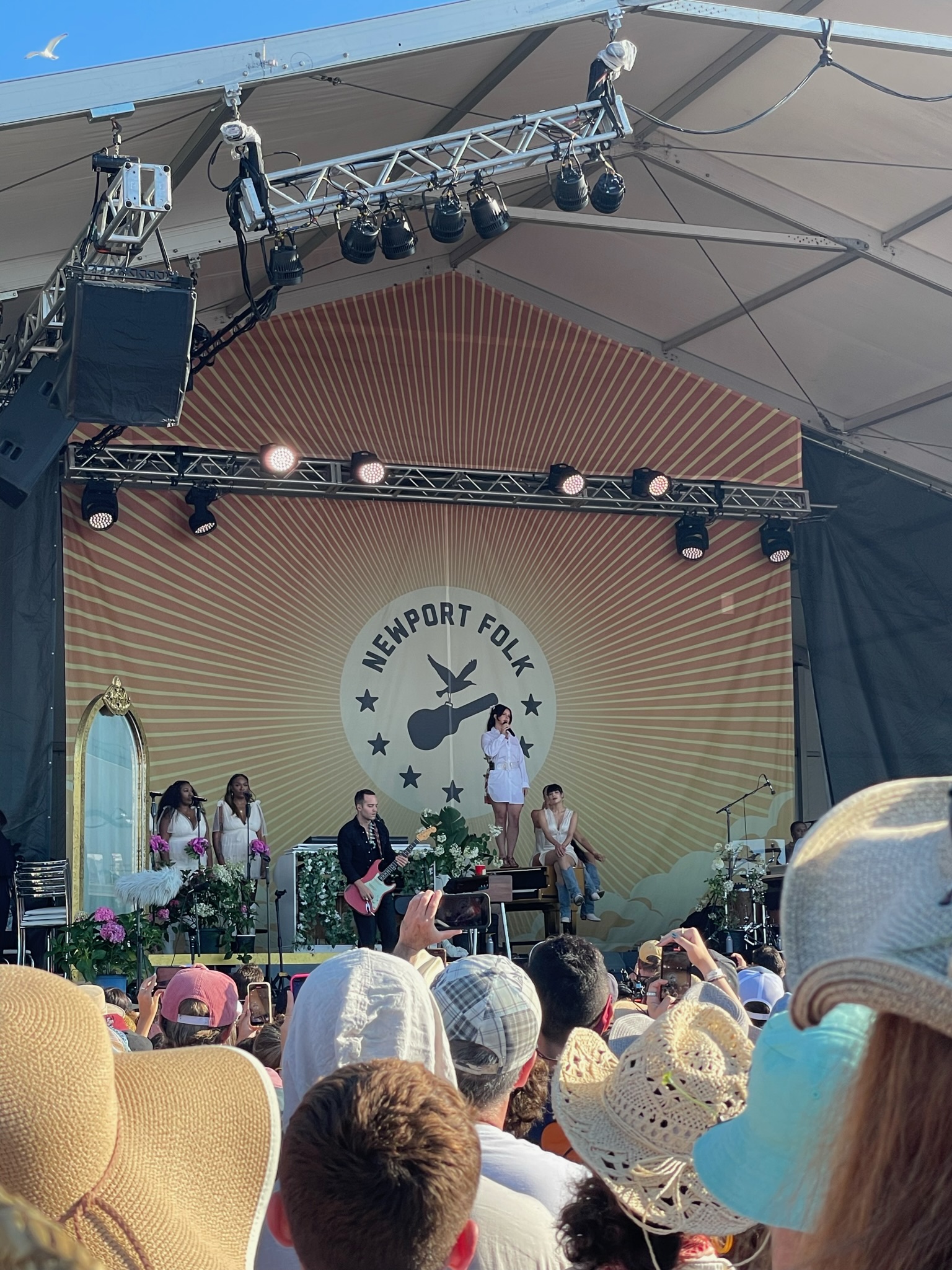 A large crowd watches a lively performance at the Newport Folk Festival. The stage backdrop features the festival's logo. Several musicians and vocalists are performing, with various instruments, plants, and decorations enhancing the stage's visual appeal.