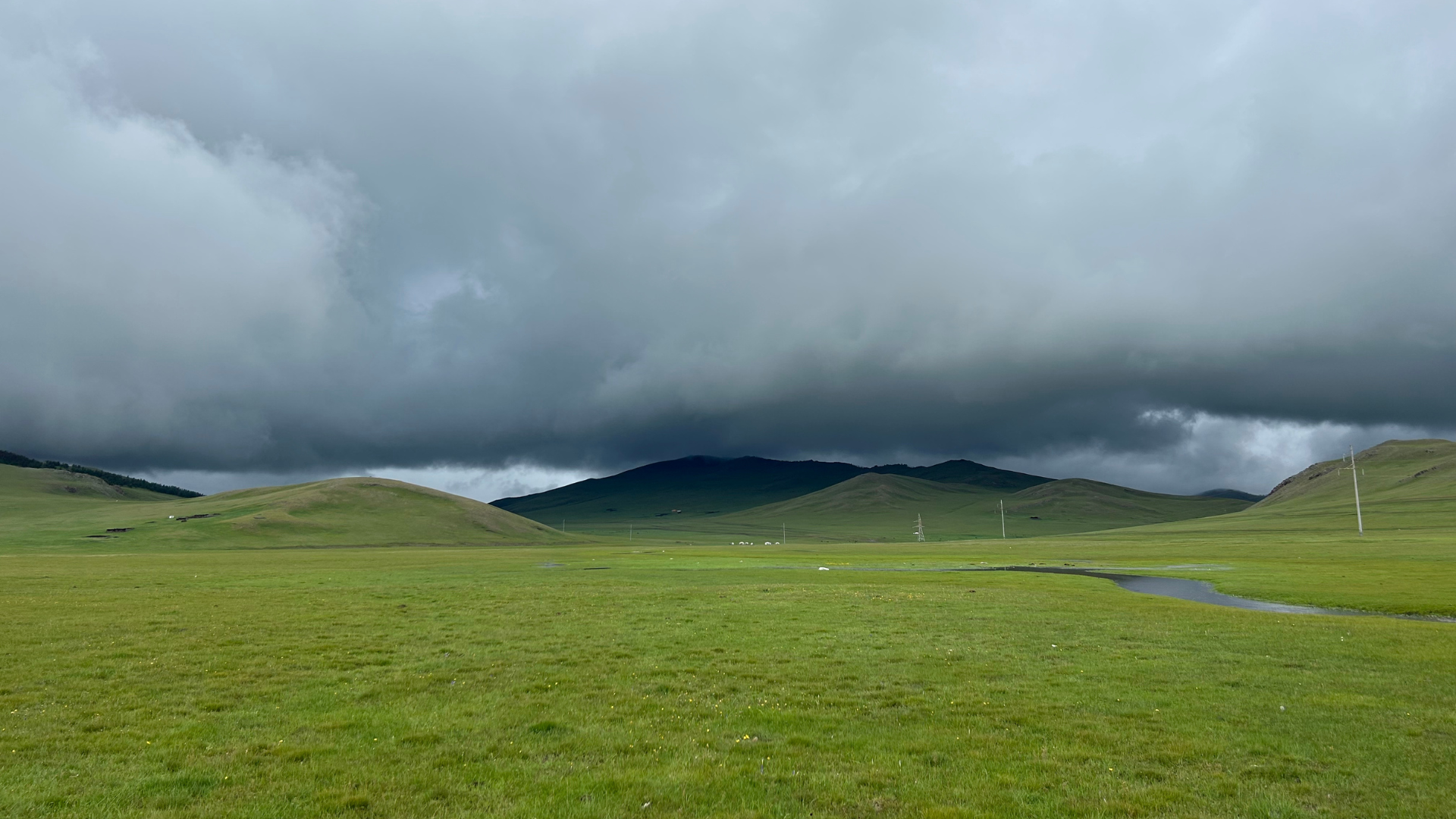 A vast green field stretches out under a dark, cloudy sky. Rolling hills rise in the background, partially shrouded in mist. A small winding stream is visible on the right side, and power lines cut across the landscape.