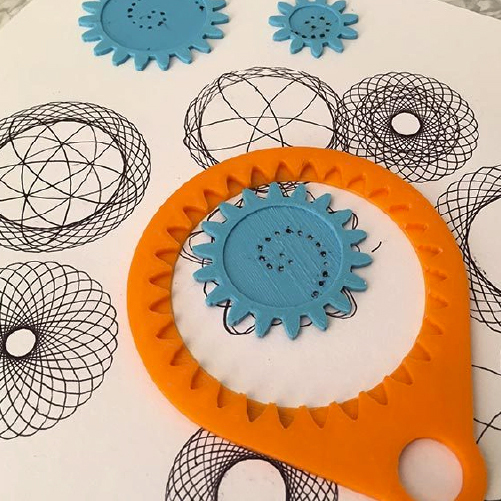 A spirograph with blue gears and an orange frame is shown alongside intricate spiral designs drawn on white paper. The gears are positioned on the paper, contributing to the creation of geometric patterns.