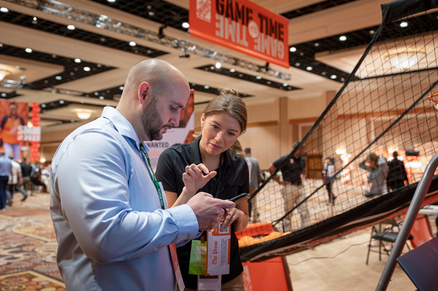 A man and a woman are engaged in a conversation at a trade show booth. The man is holding a smartphone while the woman points at it. The booth has a basketball game setup and a "GAME TIME" sign overhead. Other exhibitors and attendees are visible in the background.
