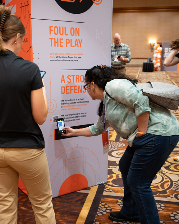 Two people, a man and a woman, are interacting with a display board in a conference or event setting. The woman is scanning a QR code with her phone, while the man observes. The background shows other attendees and event decor.