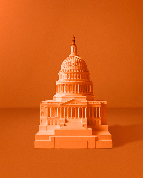 A monochromatic orange image of a model of the United States Capitol building. The model features the iconic dome, columns, and steps of the Capitol, set against a solid orange background.
