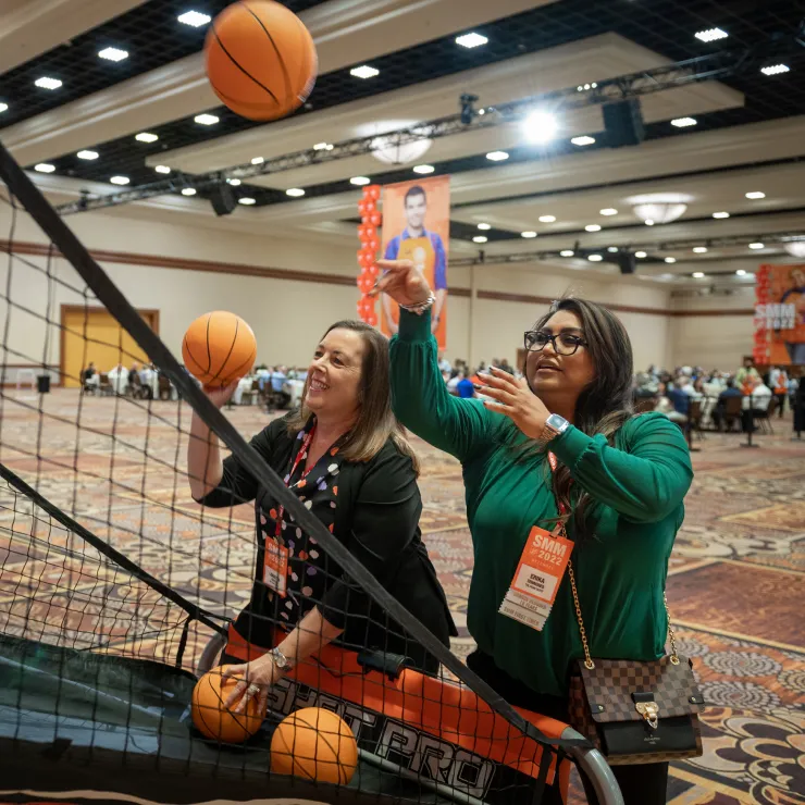 Two women are playing an arcade basketball game in a large indoor venue. Both are smiling and aiming to score. The woman on the right wears glasses and a green shirt, while the woman on the left wears a dark jacket with a name tag. Other people and event signage are in the background.
