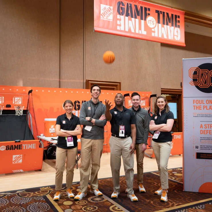 A group of five people stand under a banner that reads "Game Time" with a small basketball hoop and themed backdrop behind them. One person in the middle is tossing a basketball. They are all wearing matching uniforms with black shirts and khaki pants.