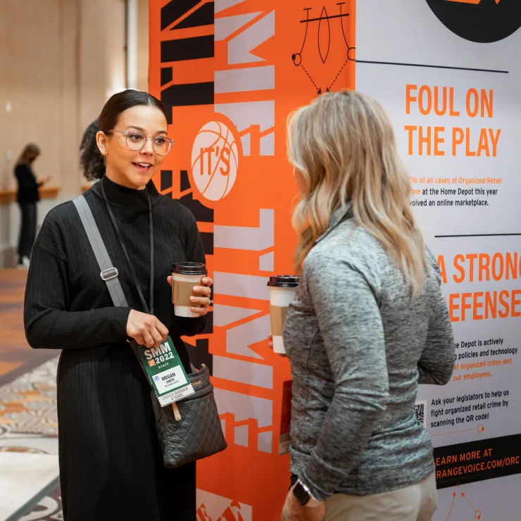 Two women are standing and talking in front of an informational display about products and technology. One is holding a coffee cup and wearing a badge with "SMI" on it. The other, facing away, also holds a coffee cup. The display has orange and white text about trade.
