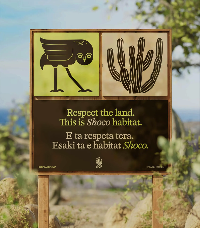 A sign in a natural area with text in English and Papiamento reads, “Respect the land. This is Shoco habitat.” The top left shows a Shoco owl, and the top right depicts a cactus. Surrounding the sign are rocks and greenery.