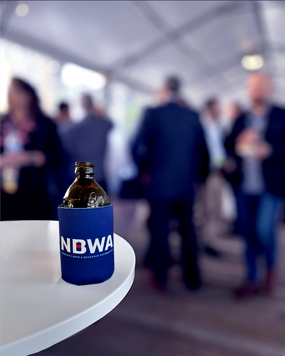 A beer in a blue koozie labeled "NDWA" rests on a white round table. The background shows a blurred crowd of people in business attire, standing and conversing in what appears to be a social event or gathering under a tent.