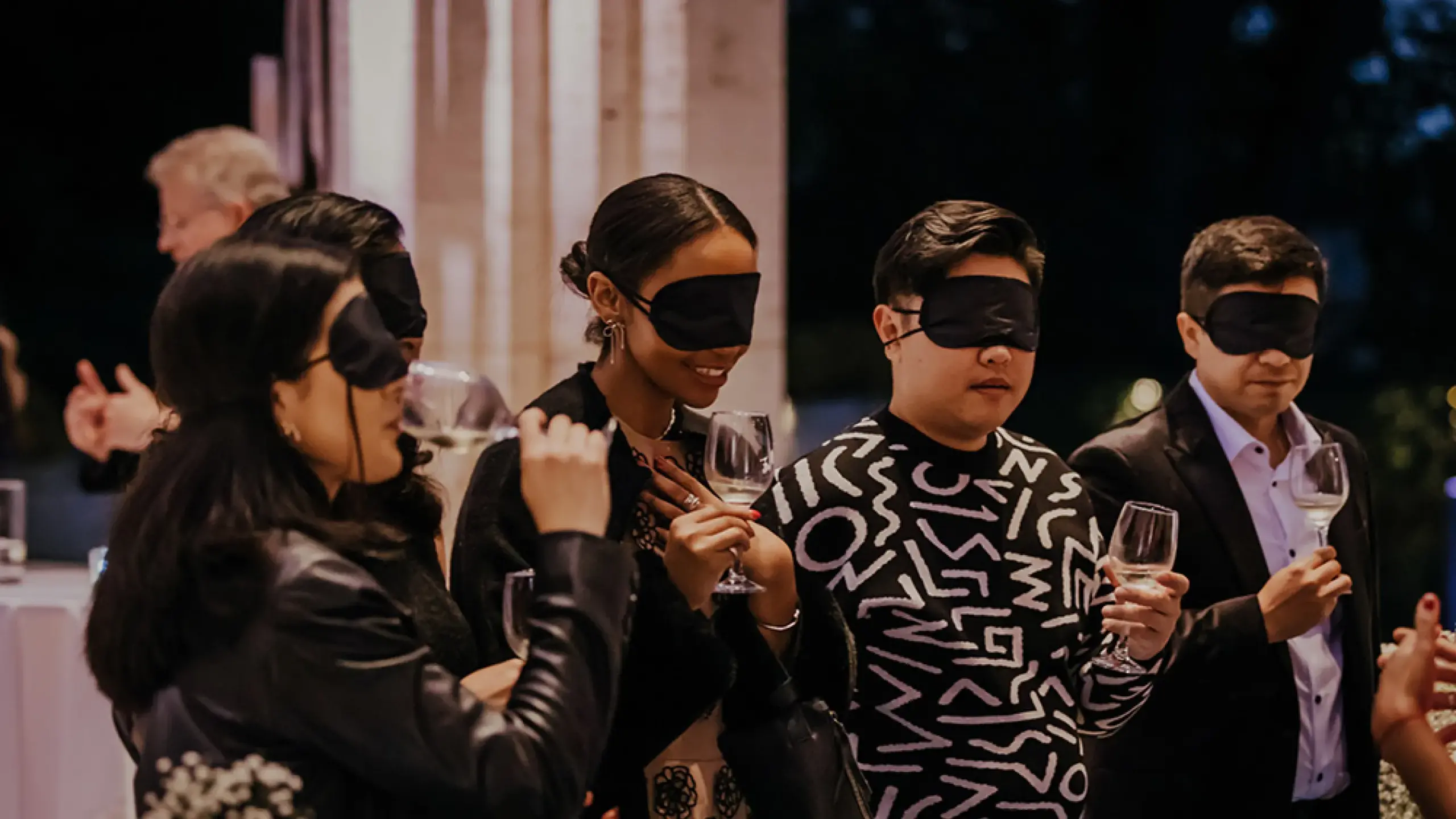 A group of people wearing black blindfolds stands together, smiling and holding wine glasses. They appear to be at a social event, engaging in what might be a sensory wine-tasting experience. The background is dimly lit with pillars and ambient lighting visible.
