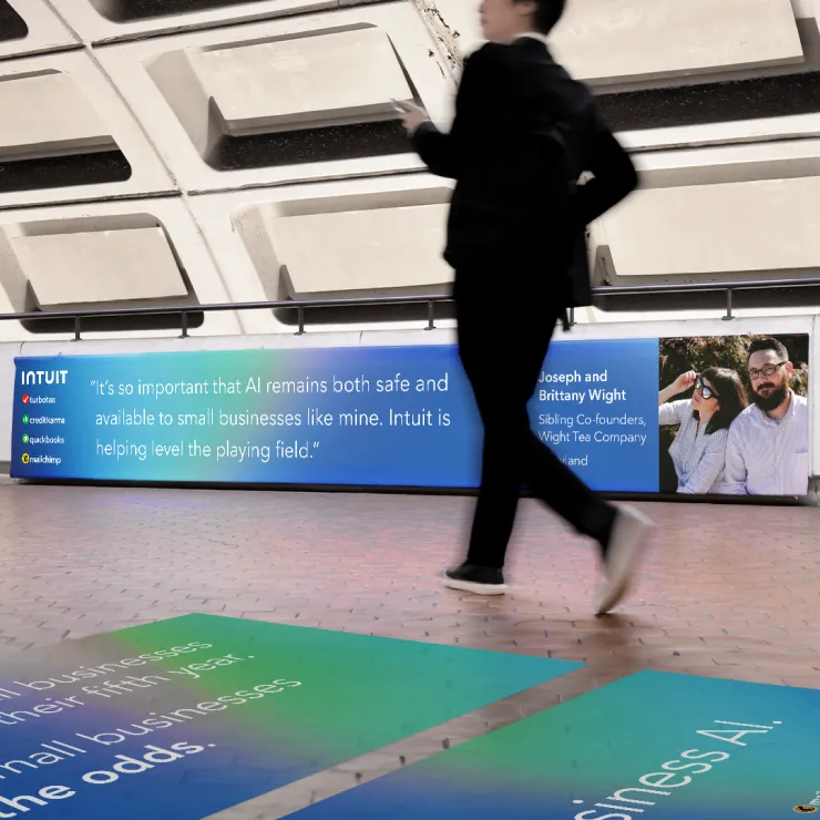 A person walks by an advertisement in a subway station. The ad has text about the importance of AI for small businesses and features images of two people, Joseph and Brittany Wight, co-founders of Wright Tea Company. The ad promotes Intuit services.