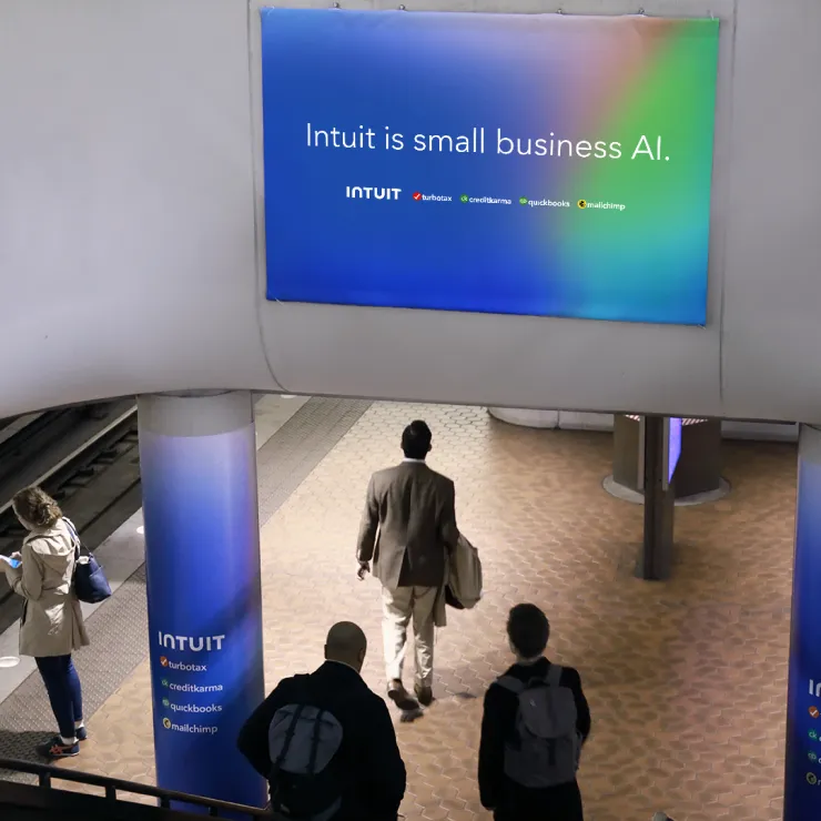 Commuters walk through a subway station with an advertisement banner above and on pillars. The large banner reads "Intuit is small business AI" and lists products like TurboTax, Credit Karma, QuickBooks, and Mailchimp.