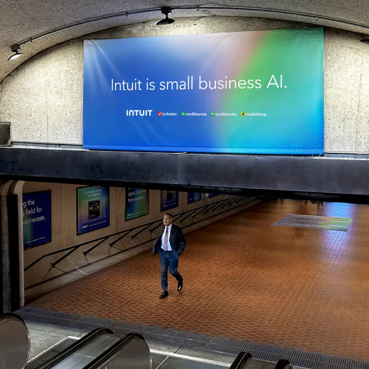 A man in formal attire walks down a tiled hallway in a subway station, passing beneath an escalator. Overhead, a large banner hangs on the wall, displaying the text "Intuit is small business AI" with logos of associated brands like Turbotax, Credit Karma, QuickBooks, and Mailchimp.