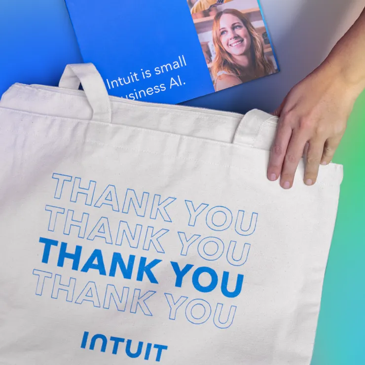 A hand is holding a white tote bag with the text "THANK YOU THANK YOU THANK YOU THANK YOU" and the Intuit logo on it. Behind the bag is a blue booklet featuring a smiling person and the text "Intuit is small business AI." The background has a gradient color effect.