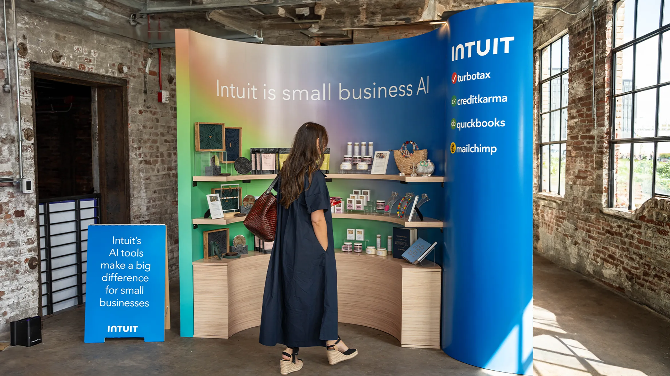 A person stands in front of an Intuit display booth with a colorful backdrop that reads, "Intuit is small business AI." The booth features various products and promotional materials. A sign next to the booth reads, "Intuit's AI tools make a big difference for small businesses.