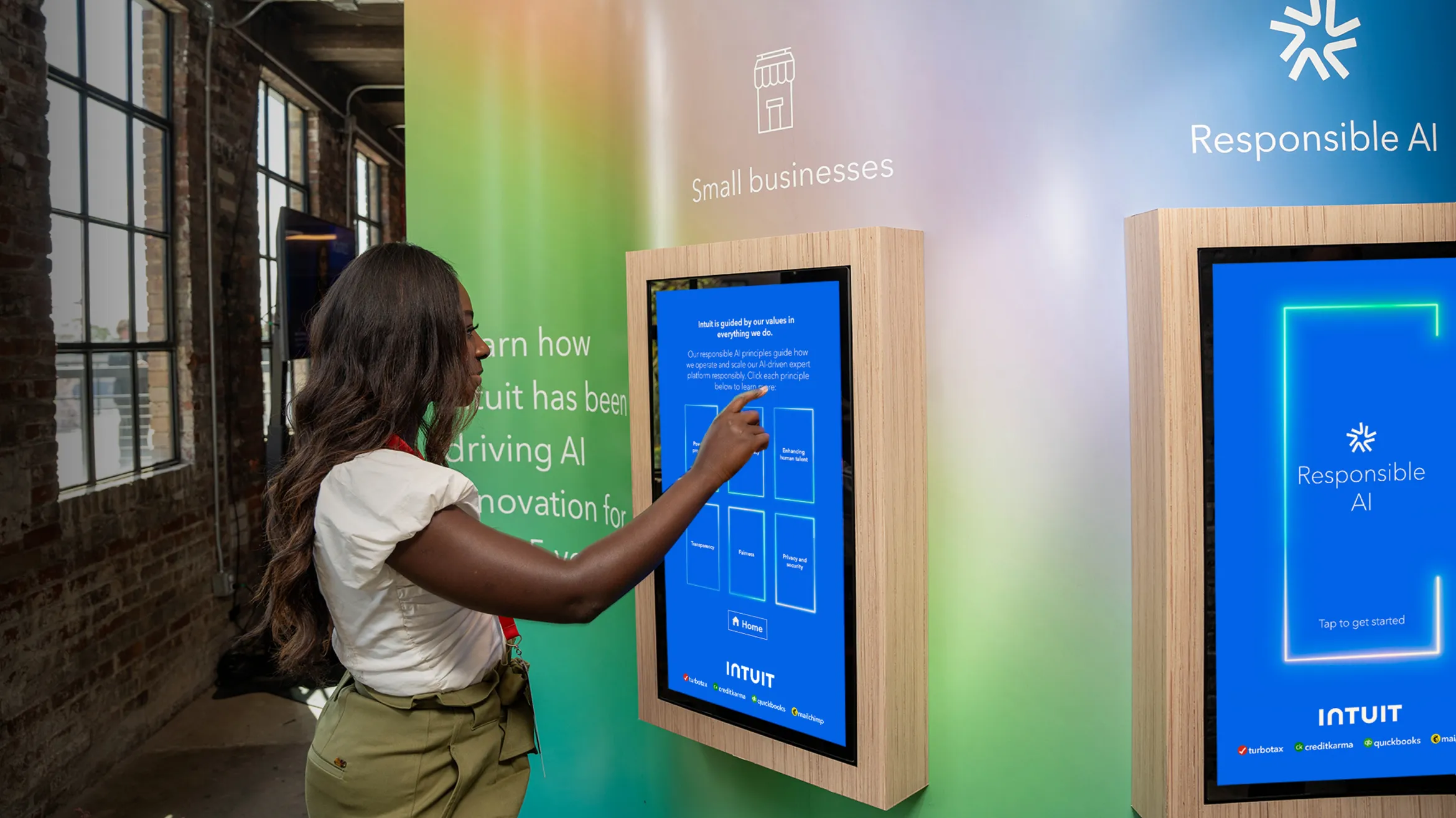 A person with long hair interacts with a digital information kiosk displaying content about small businesses and responsible AI. The interface is vibrant with a blue screen, and the surroundings have soft, colorful backdrops and wooden frames.