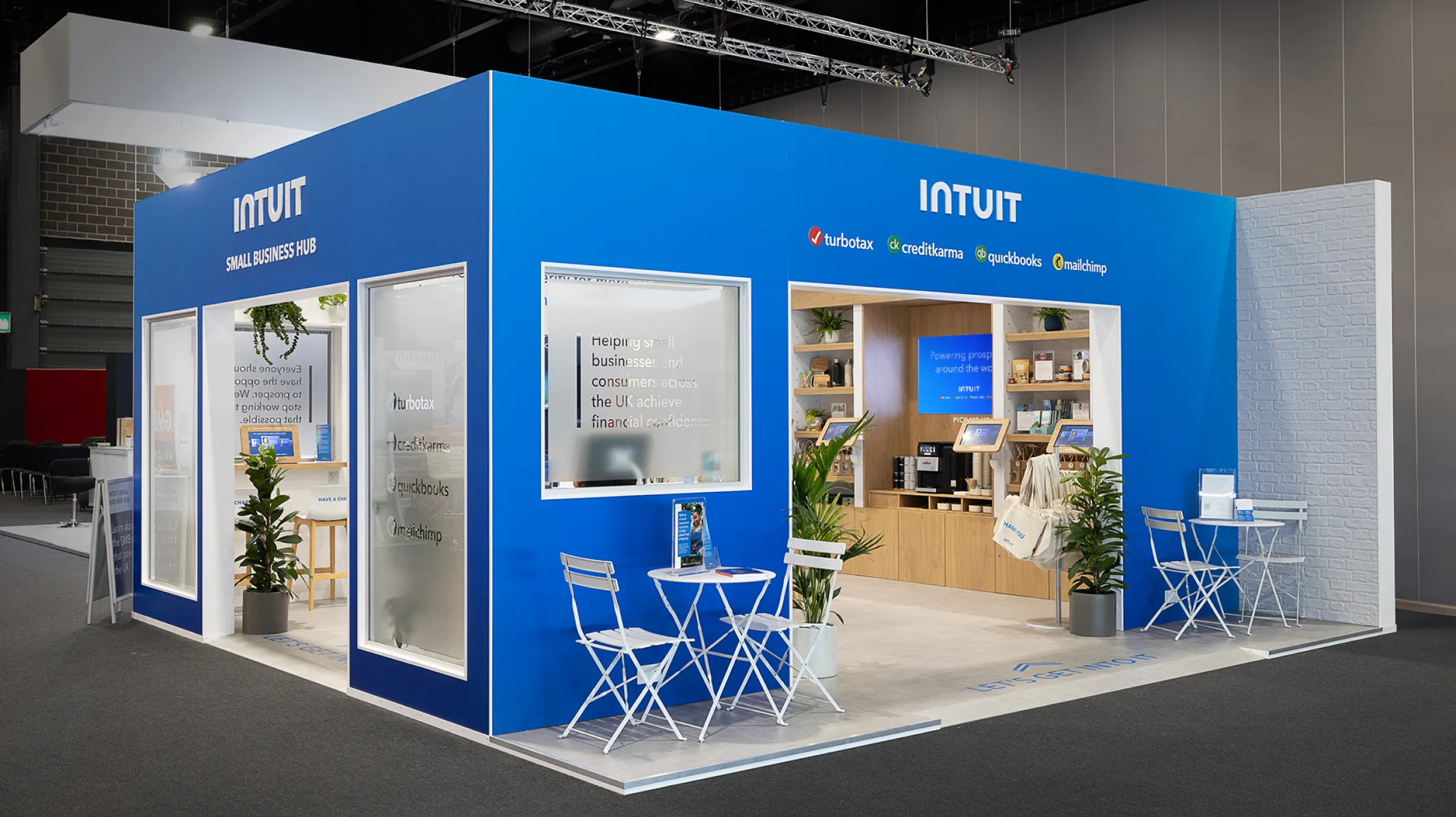 A blue booth at a trade show labeled "INTUIT," featuring the logos "Turbotax," "Creditkarma," "Quickbooks," and "Mailchimp." The booth has glass windows, potted plants, small white tables and chairs, and promotional materials displayed inside.