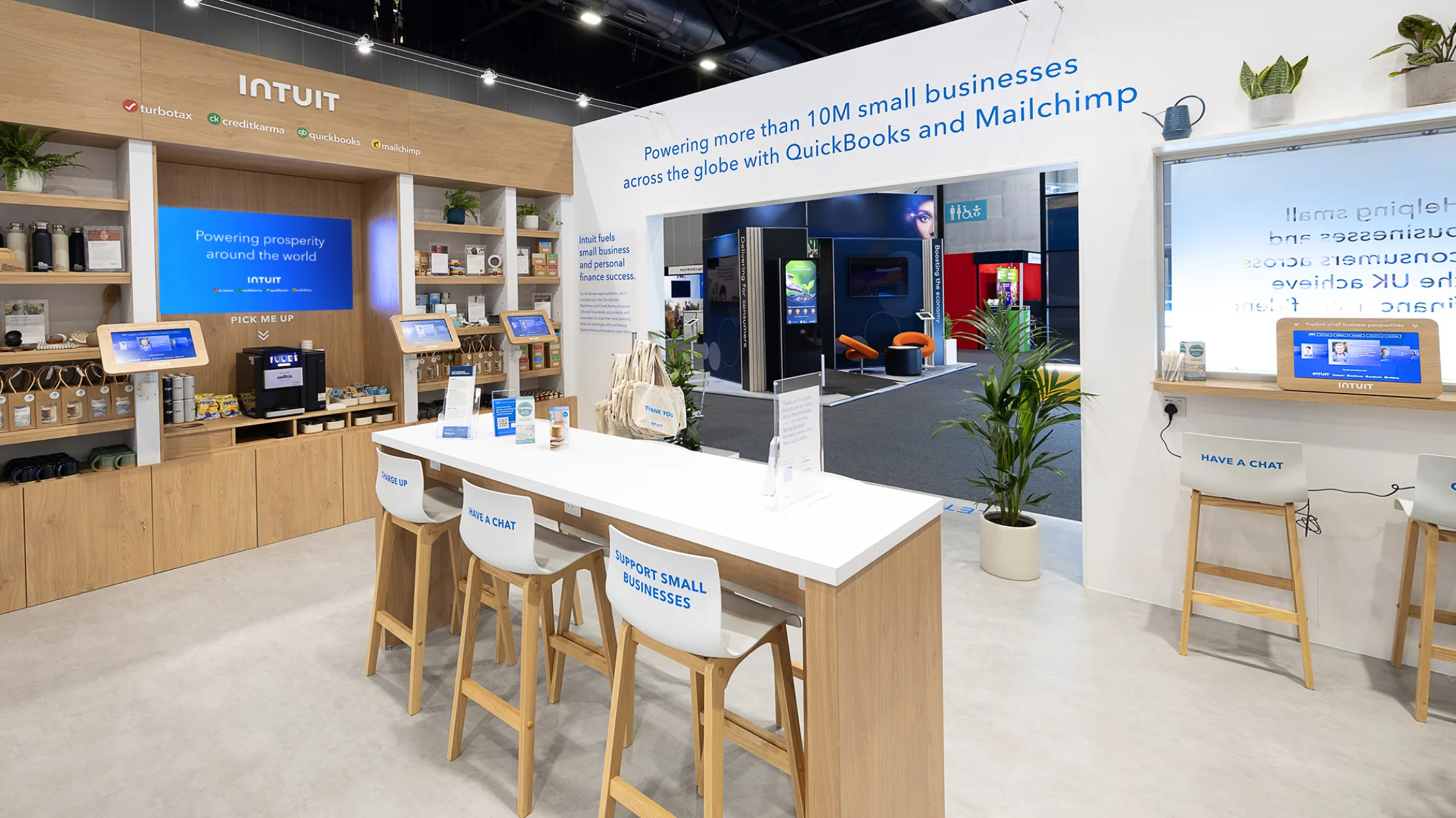 A trade show booth for Intuit, promoting QuickBooks and Mailchimp. The booth features a long counter with stools, various display screens, and promotional materials. The backdrop has the slogan "Powering more than 10M small businesses across the globe.