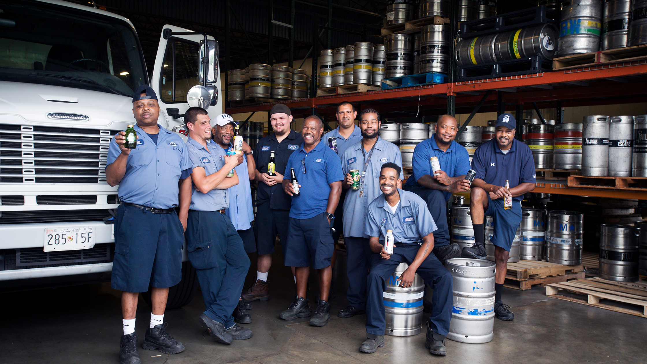 A group of eleven men in blue work uniforms pose with drinks in front of a delivery truck and stacked metal kegs in a warehouse. They appear relaxed and are smiling, some leaning on kegs or the truck.