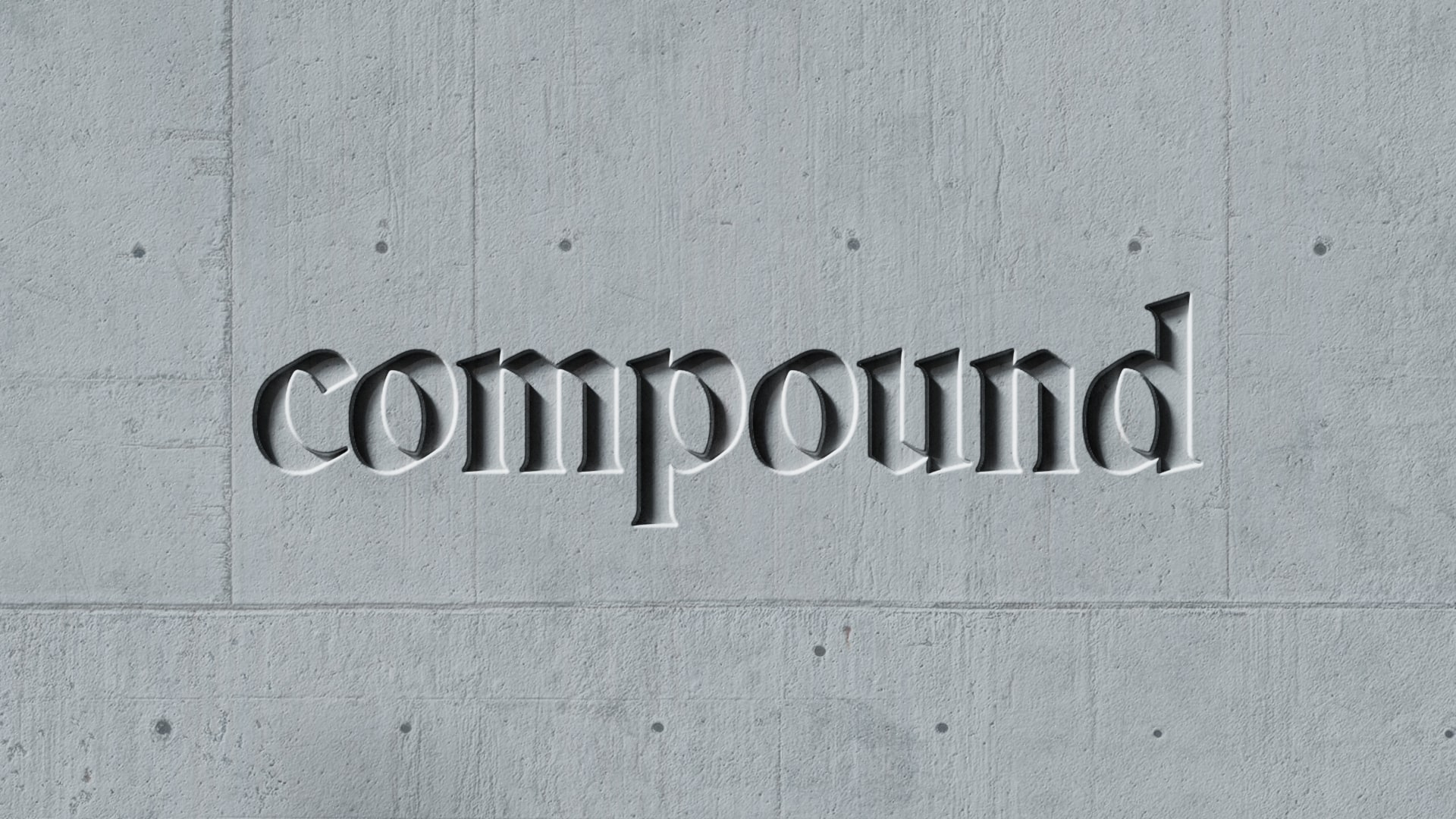 The word "compound" is chiseled into a smooth, gray concrete wall, creating a 3D effect with shadows and highlights that give the text depth. The background is slightly textured but uniform in color.