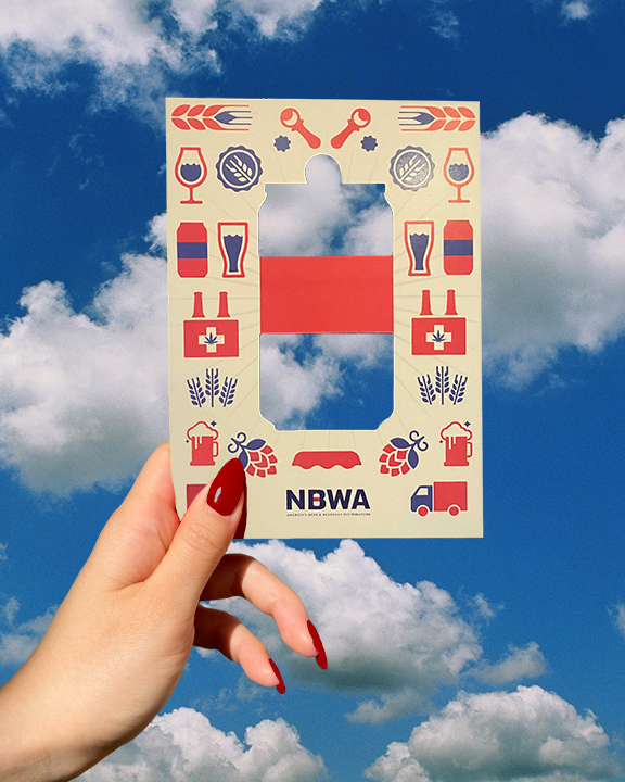A hand with red-painted nails holds up a beige stencil with various red and blue beer-themed icons, such as mugs, bottles, wheat, and tools, against a backdrop of a blue sky with fluffy white clouds. The stencil has a cut-out section and "NBWA" written at the bottom.