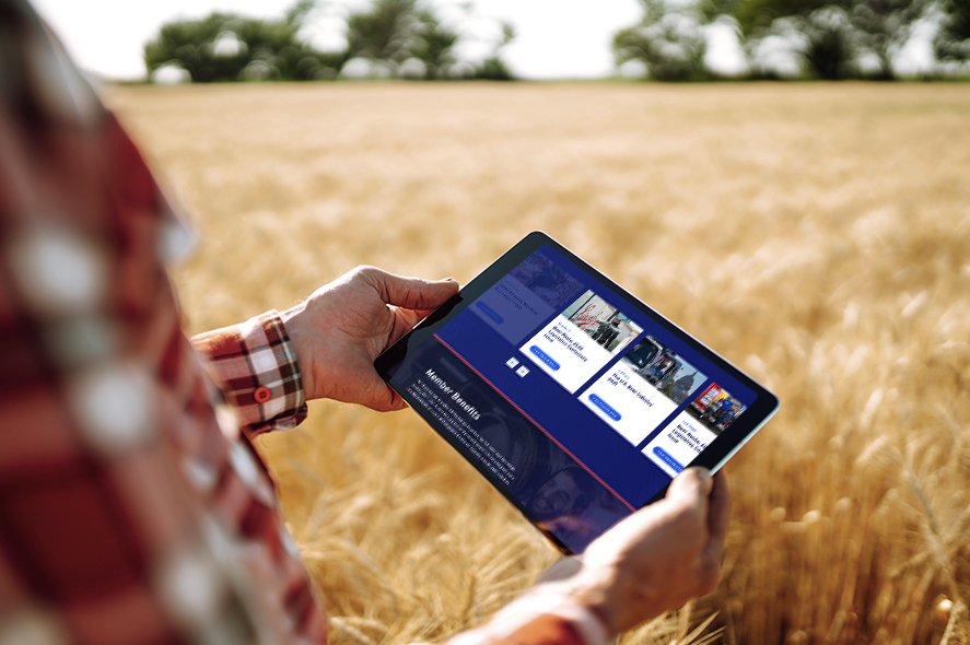 A person in a plaid shirt holds a tablet in a wheat field. The tablet's screen displays an agricultural app with images of crops and options like "View Report" and "Soil Analysis." The background shows a clear sky and green trees.