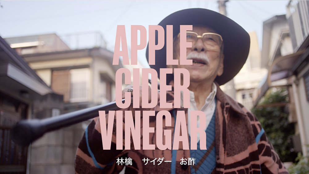 An elderly man wearing a hat and glasses stands outdoors, smiling and holding a cane. The text "APPLE CIDER VINEGAR" is prominently displayed in large pink letters across the image. Japanese text appears at the bottom. The background shows a street with buildings.