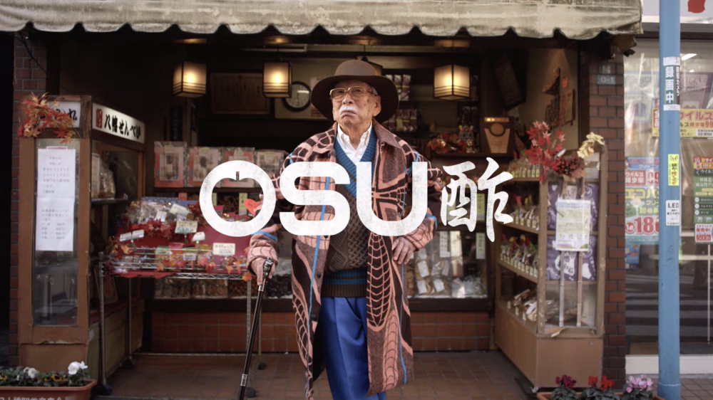 An elderly man stands confidently in front of a shop filled with various goods. He wears a hat, a patterned coat, and carries a cane. The shop has a traditional look with multiple items on display and some signage. The word "OSU" is prominently written over the image.
